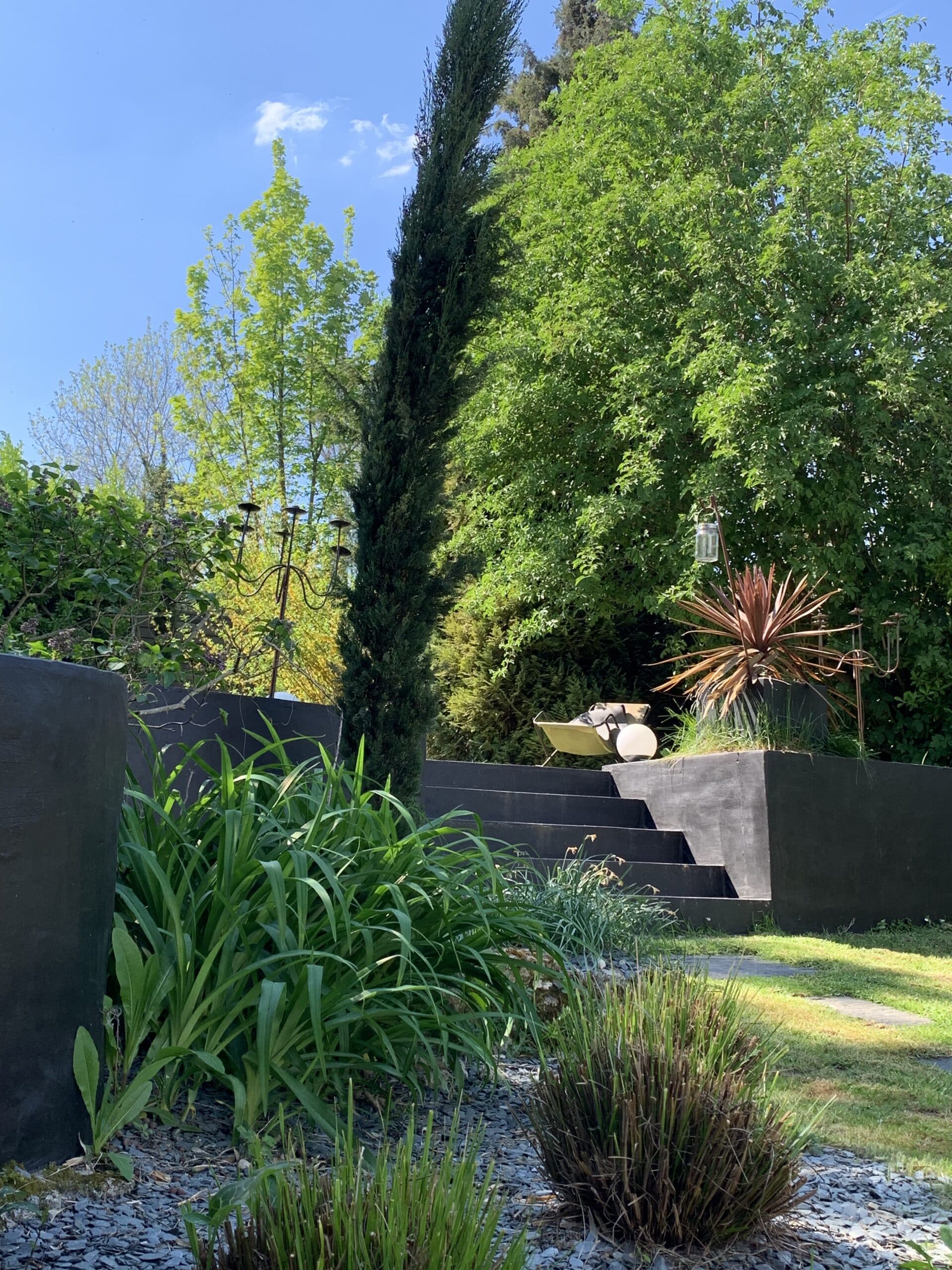 Jardin paysager moderne avec escalier noir, plantes vertes luxuriantes, un cyprès et un ciel bleu éclatant sous le soleil.