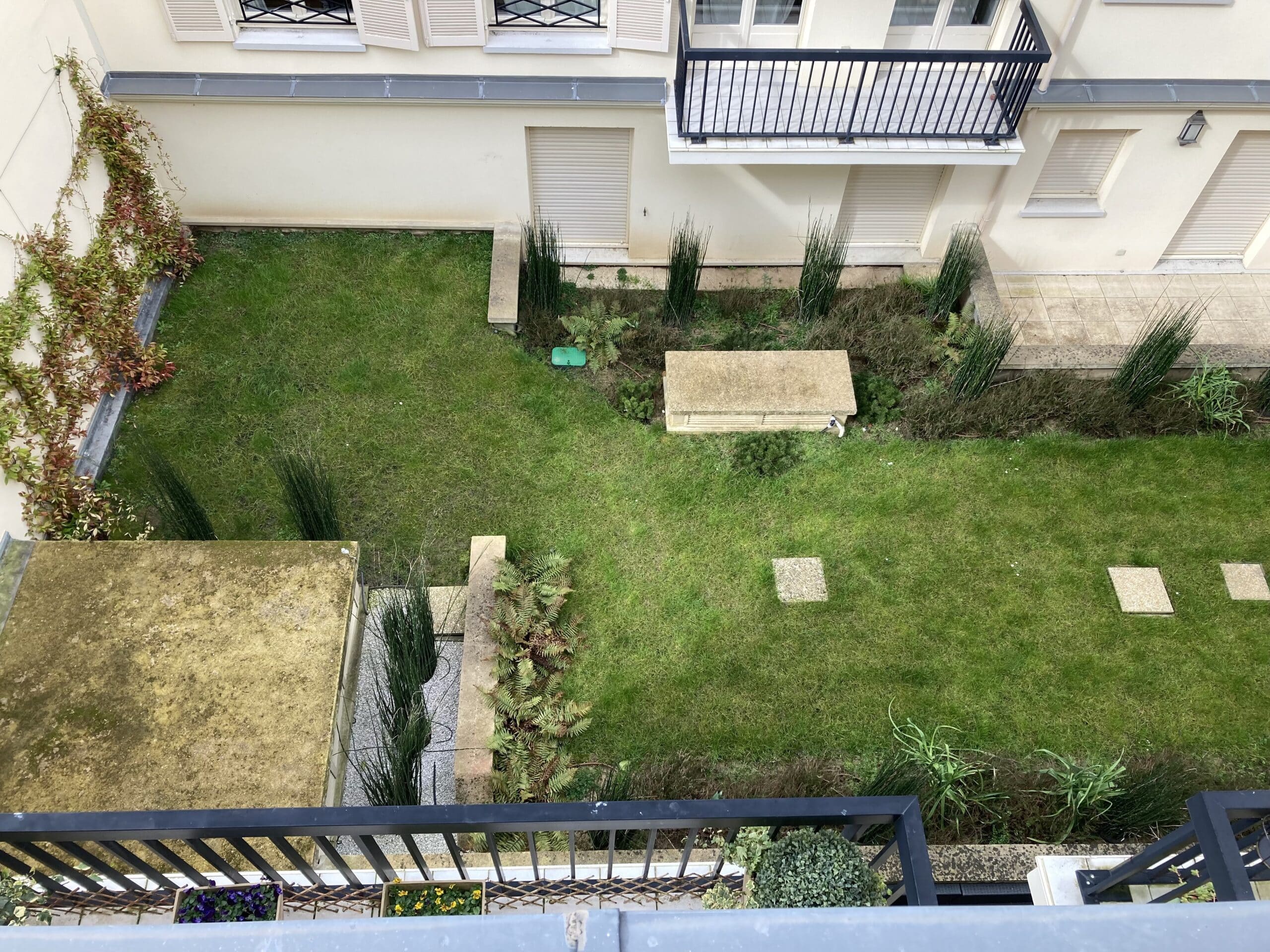 Vue aérienne d'un jardin résidentiel avec pelouse verte, plantes variées, murs crème, fenêtres à volets, et un balcon avec rambarde noire.