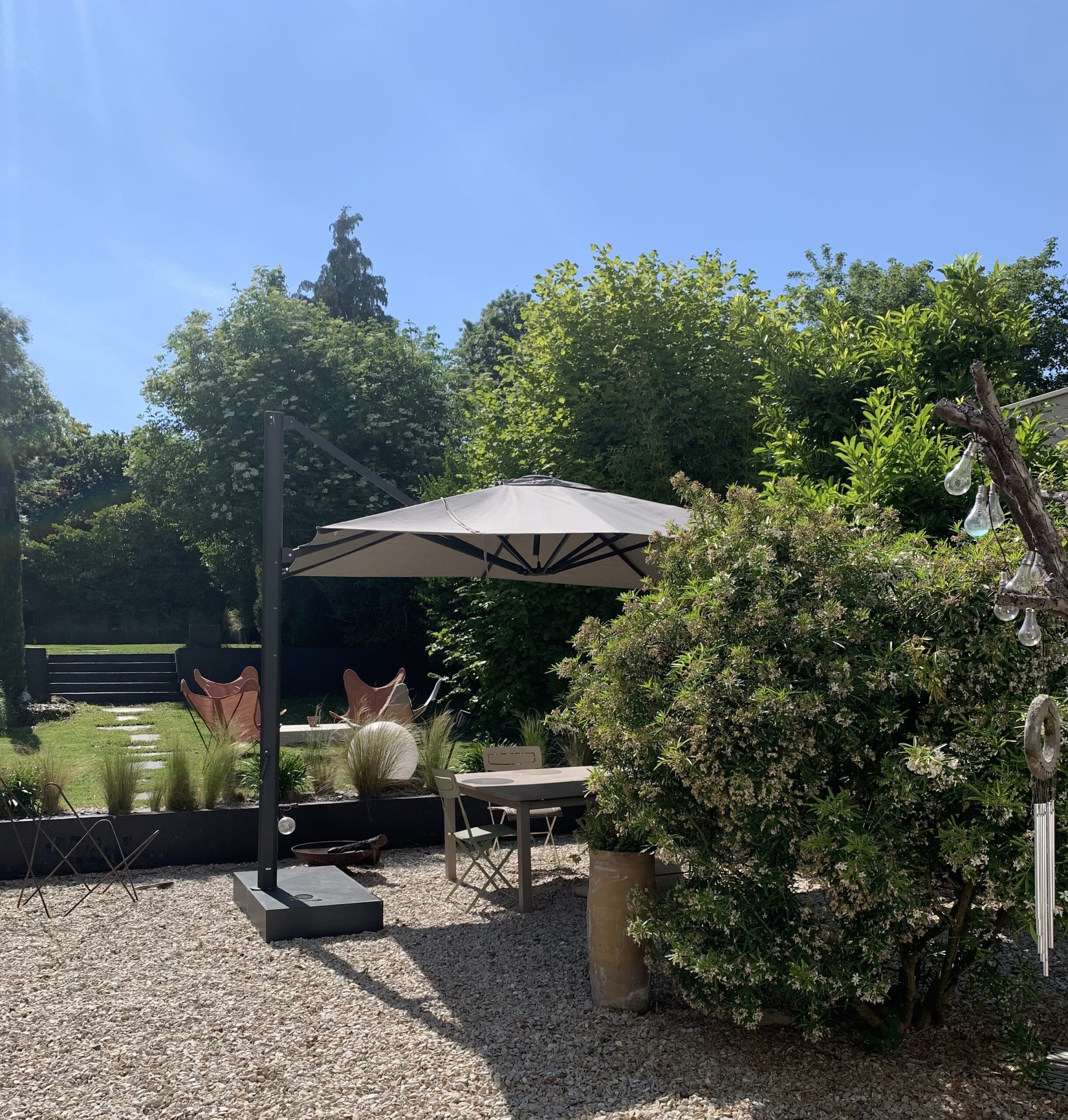 Terrasse en gravier, parasol gris, table et chaises de jardin sous un ciel bleu ensoleillé. Verdure luxuriante et coin détente avec fauteuils.