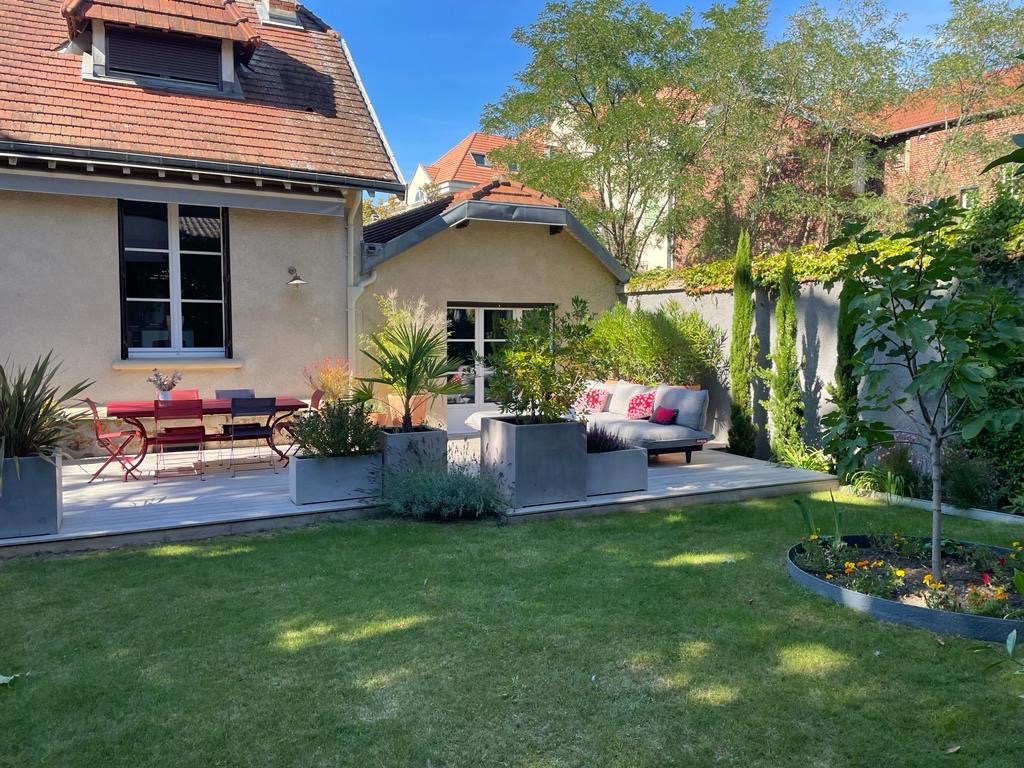 Jardin aménagé avec terrasse en bois, salon de jardin gris et table rouge. Gazon vert, plantes en pots et arbres encadrent la maison ensoleillée.