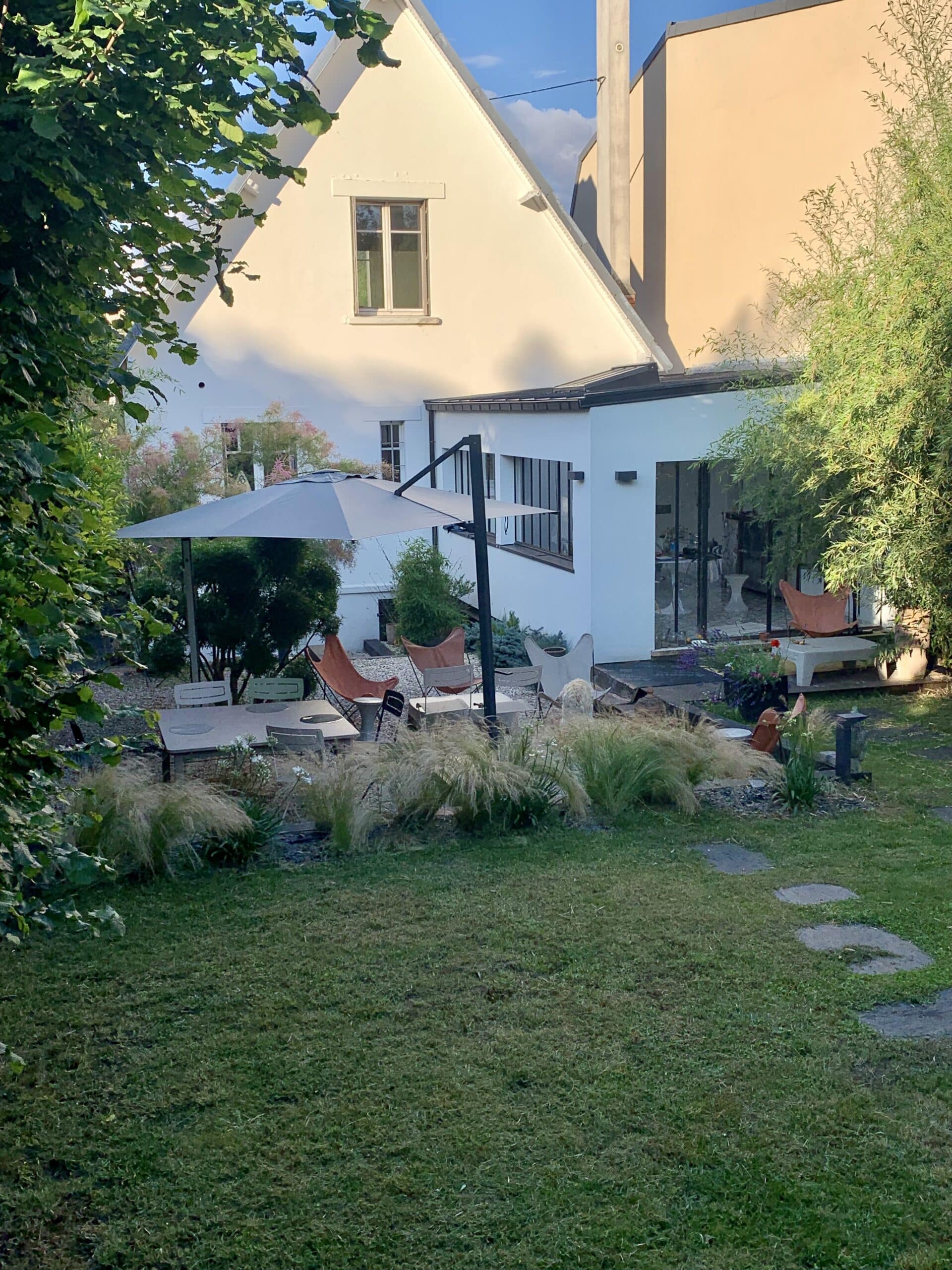 Jardin verdoyant avec terrasse moderne, parasol, mobilier design et une maison contemporaine en arrière-plan sous un ciel bleu.