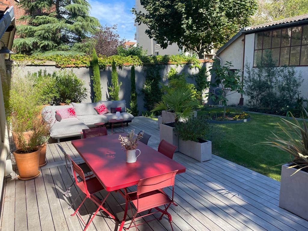Terrasse en bois ensoleillée avec salon de jardin gris et coussins rouges. Table à manger rouge et chaises, entourée de plantes et d'un jardin.