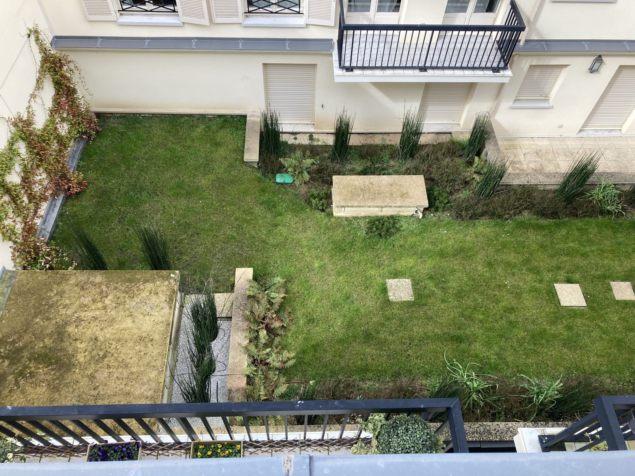 Vue plongeante sur un jardin urbain avec pelouse verte, plantes grimpantes, balcon, et jardinières fleuries.