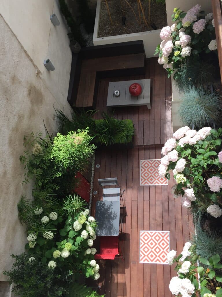 Terrasse moderne en bois. Hydrangeas blancs/roses, plantes vertes luxuriantes. Mobilier gris et rouge, banquette intégrée et motifs géométriques orange.