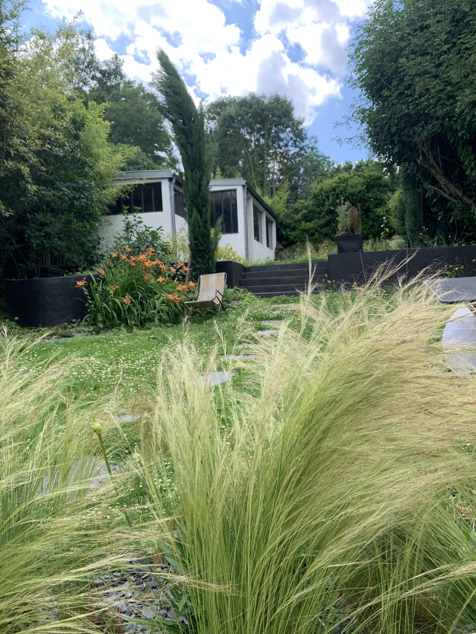 Jardin moderne luxuriant avec herbe ornementale au premier plan. Des lys oranges fleurissent devant une maison blanche, des escaliers foncés, et un ciel nuageux.