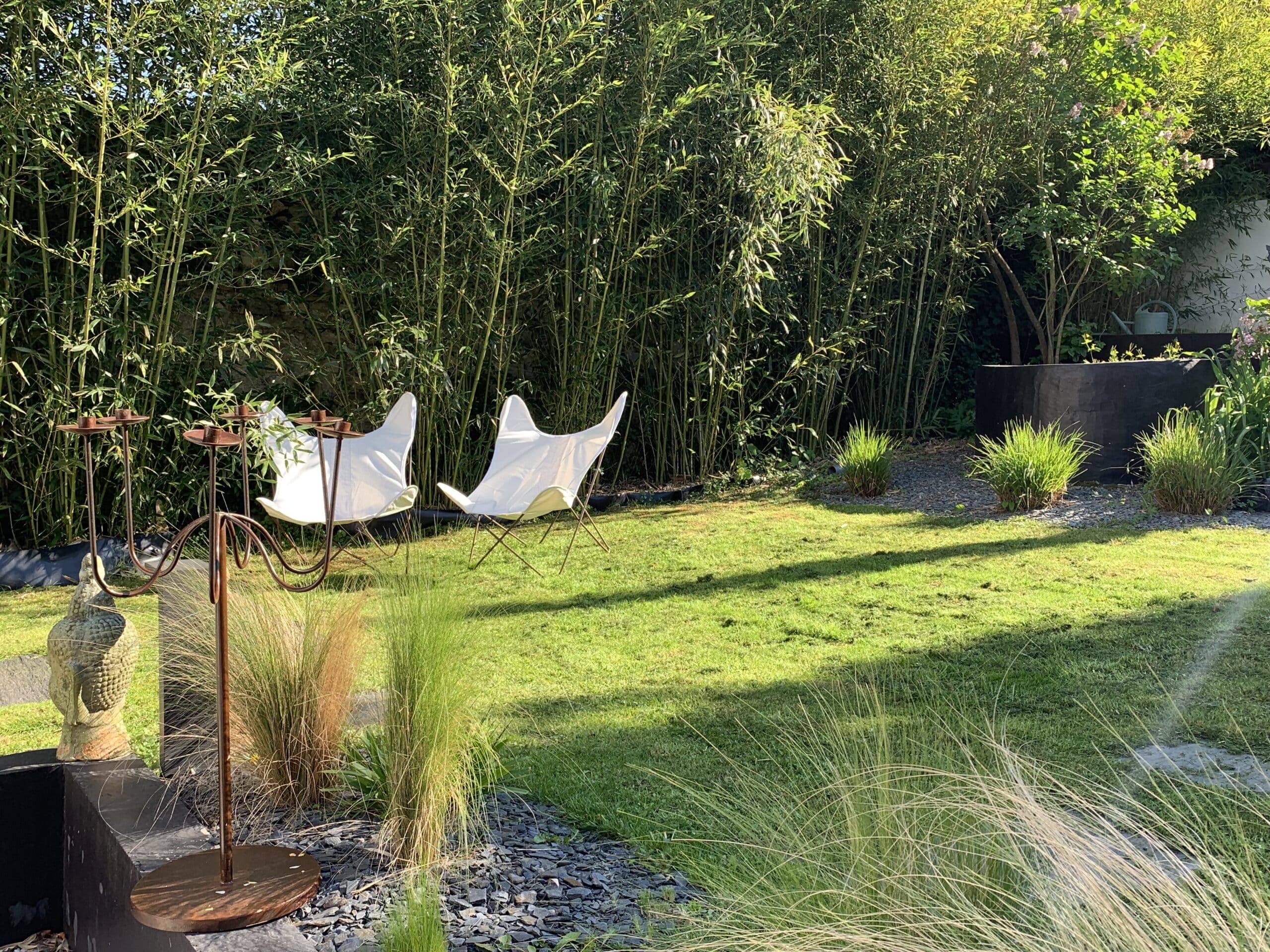 Jardin ensoleillé avec une haute haie de bambous, deux chaises papillon blanches sur la pelouse verte et un bouddha en pierre. Ambiance zen.