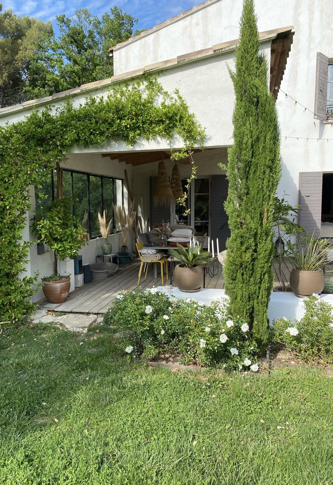 Maison blanche méditerranéenne avec terrasse boisée, vigne grimpante, cyprès élancé, et rosiers blancs. Ambiance extérieure ensoleillée.