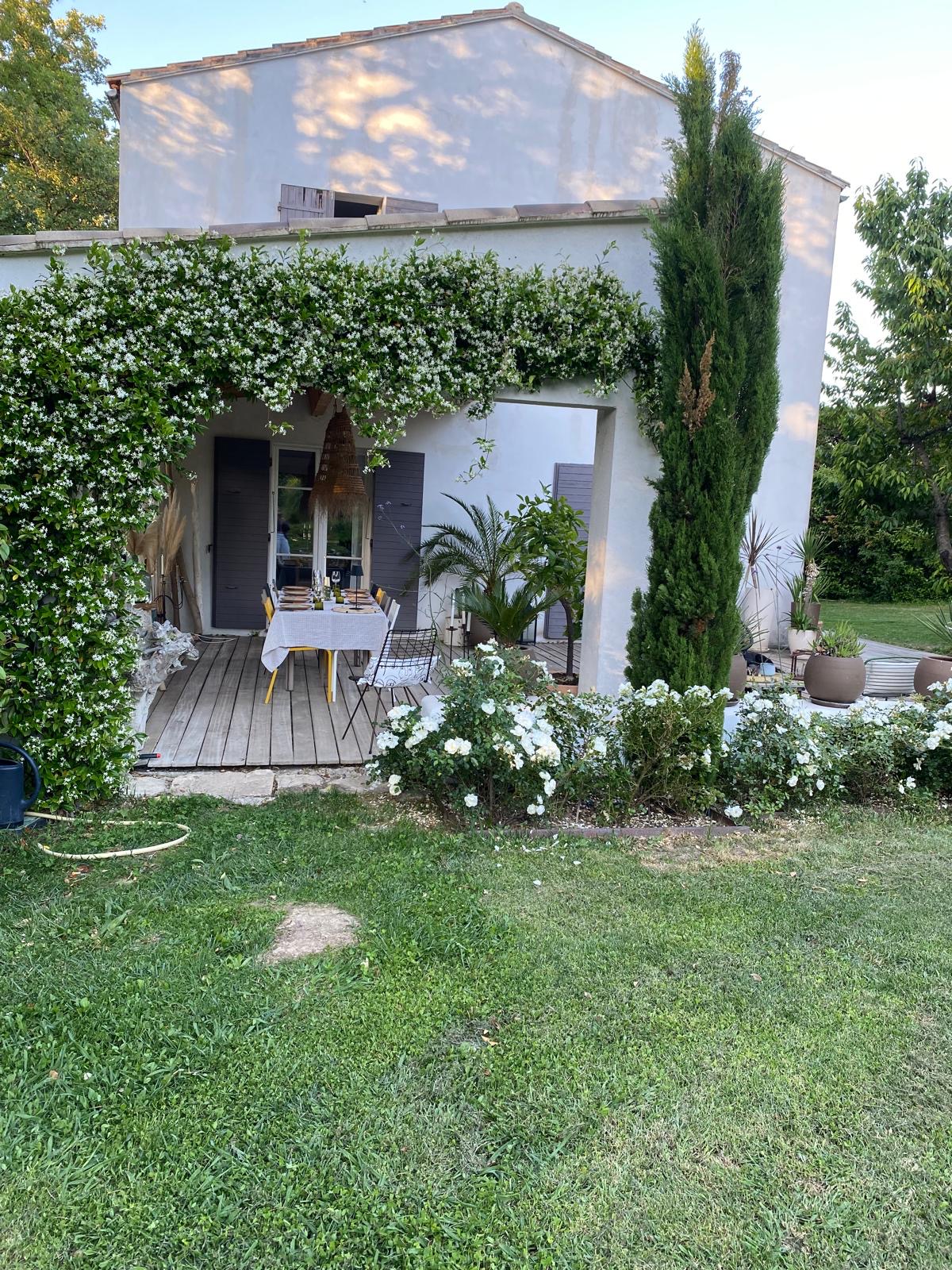 Maison blanche provençale avec patio en bois, jasmin fleuri en pergola. Table dressée pour un dîner extérieur, bordée de cyprès et rosiers blancs.