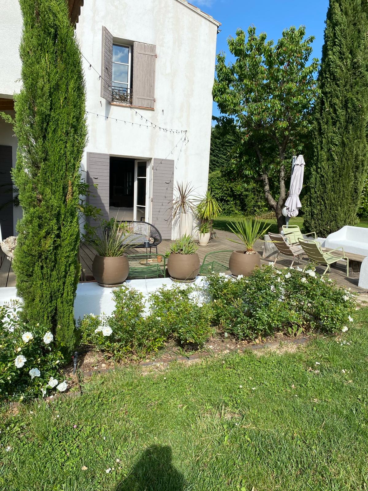 Maison blanche aux volets gris, terrasse en bois avec mobilier d'extérieur, plantes en pots et jardin de roses blanches sous ciel bleu.