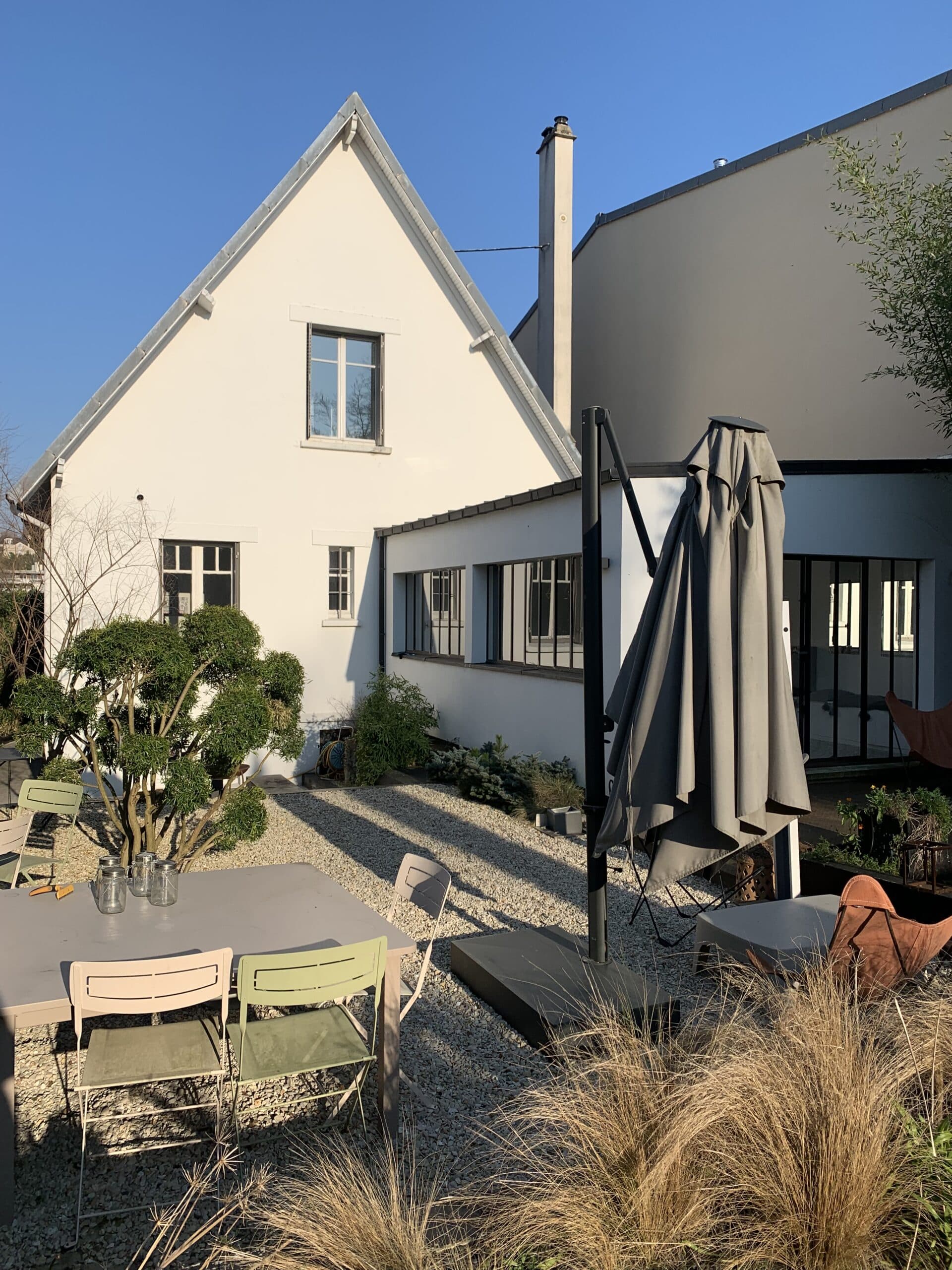 Terrasse de jardin ensoleillée avec maison blanche, sol en gravier, mobilier extérieur coloré, parasol fermé et plantes vertes.