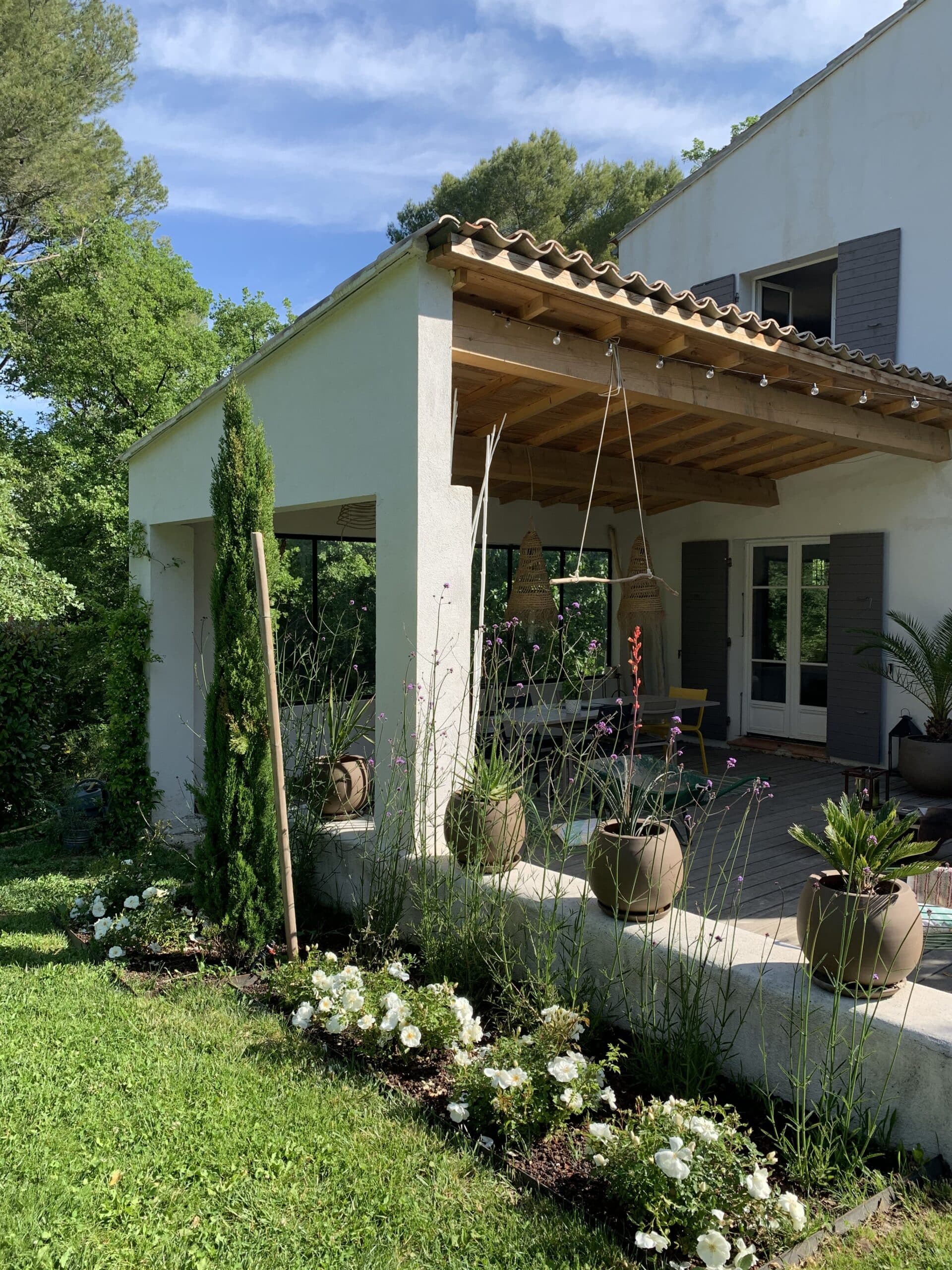 Maison provençale avec patio couvert en bois, jardin ensoleillé, gazon vert, roses blanches et cyprès. Toit en tuiles.