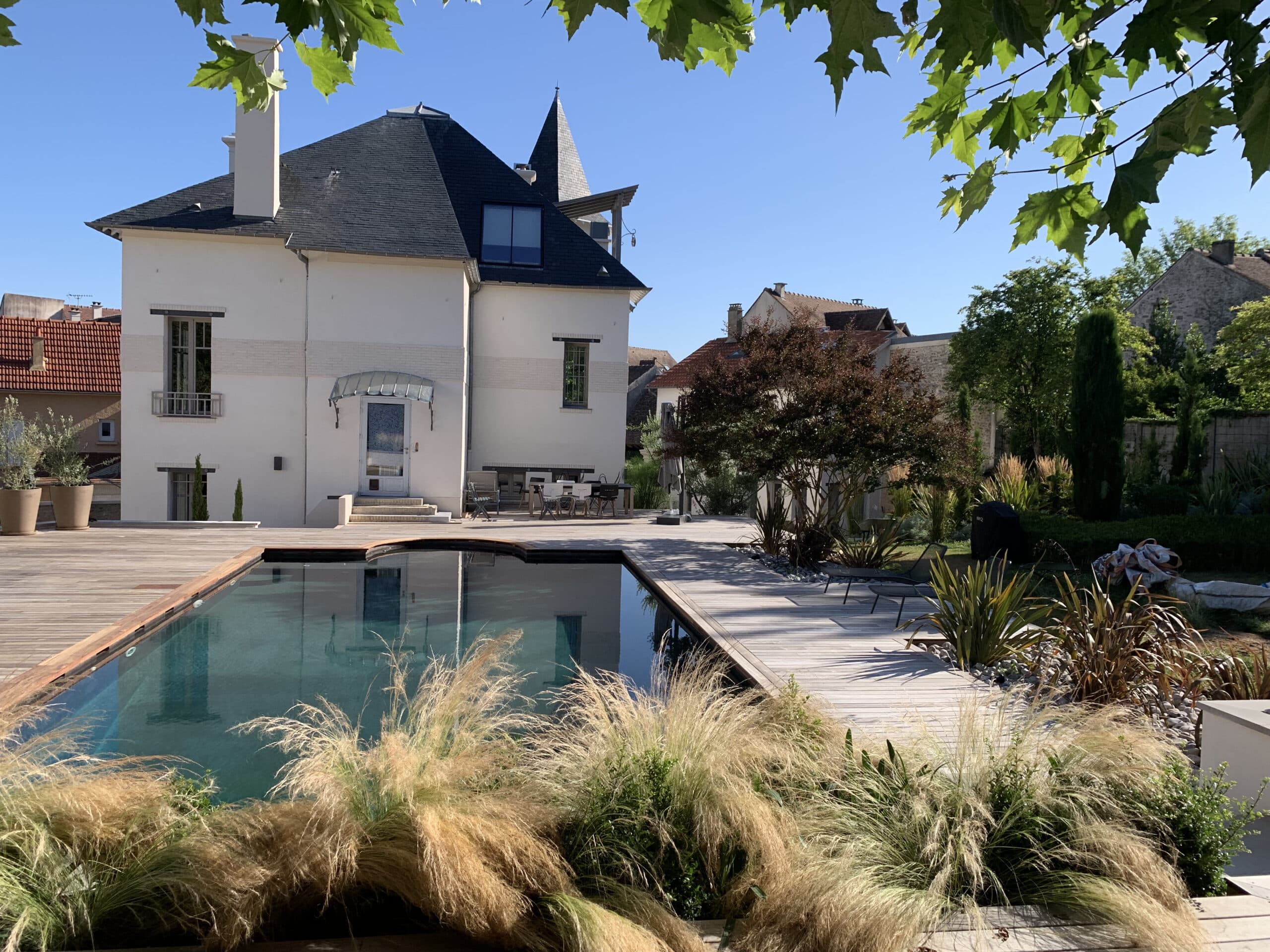 Maison élégante avec piscine rectangulaire, grande terrasse en bois clair. Jardin luxuriant de graminées, arbres verts sous un ciel bleu ensoleillé.