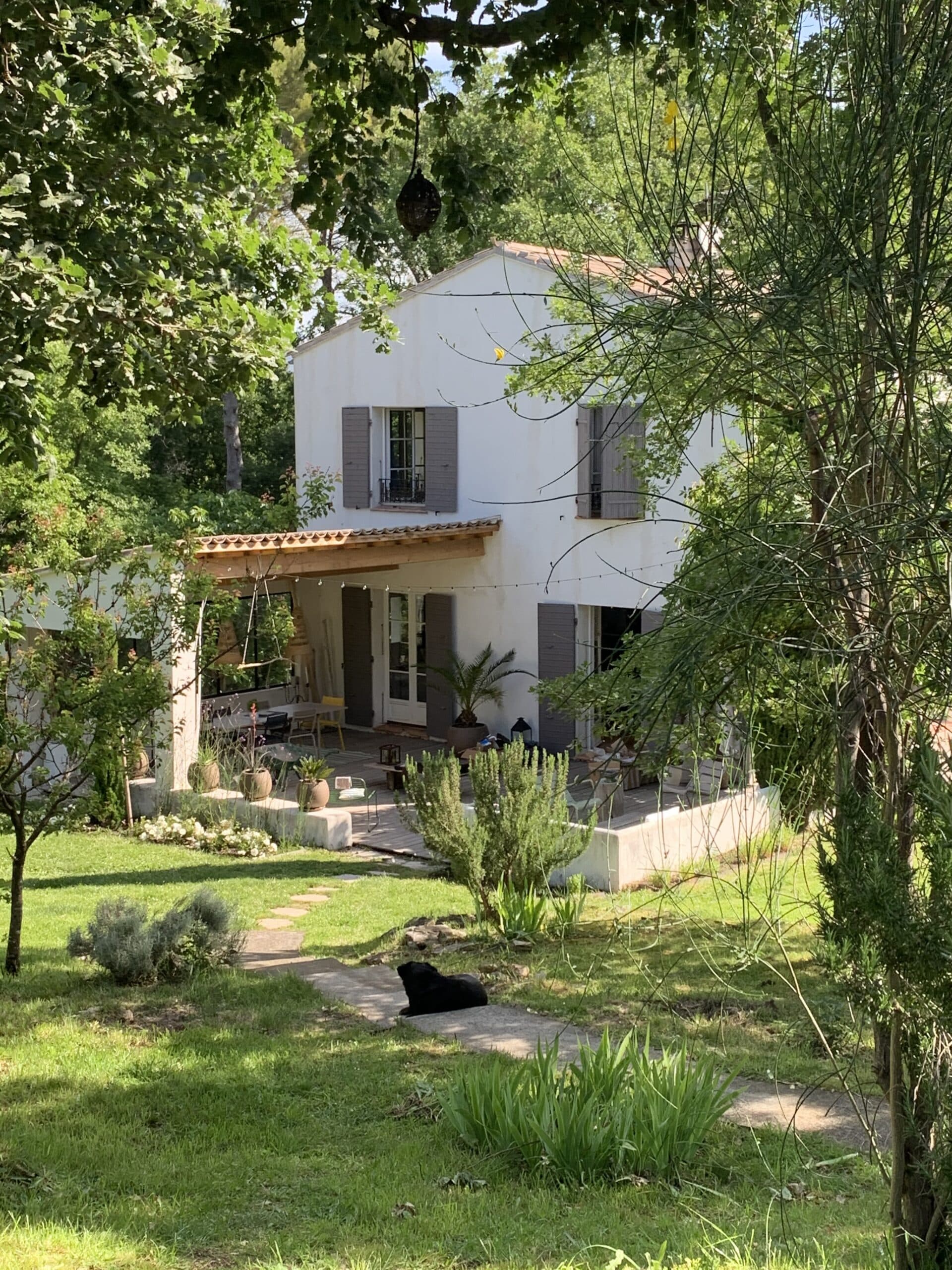 Maison blanche aux volets gris, entourée d'arbres luxuriants, avec un chien noir allongé sur un chemin dans un jardin verdoyant et ensoleillé.