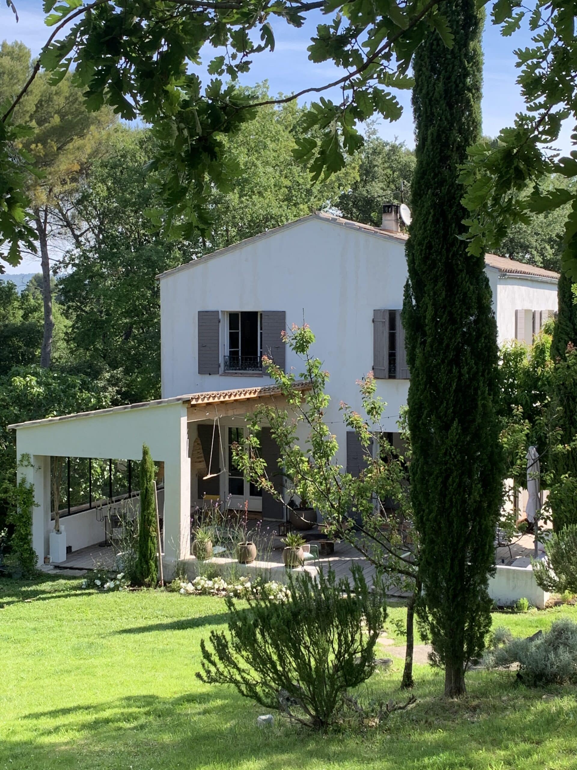 Maison blanche provençale ensoleillée avec volets marron, jardin verdoyant, arbres et cyprès élancés. Terrasse couverte avec balançoire, pelouse luxuriante.