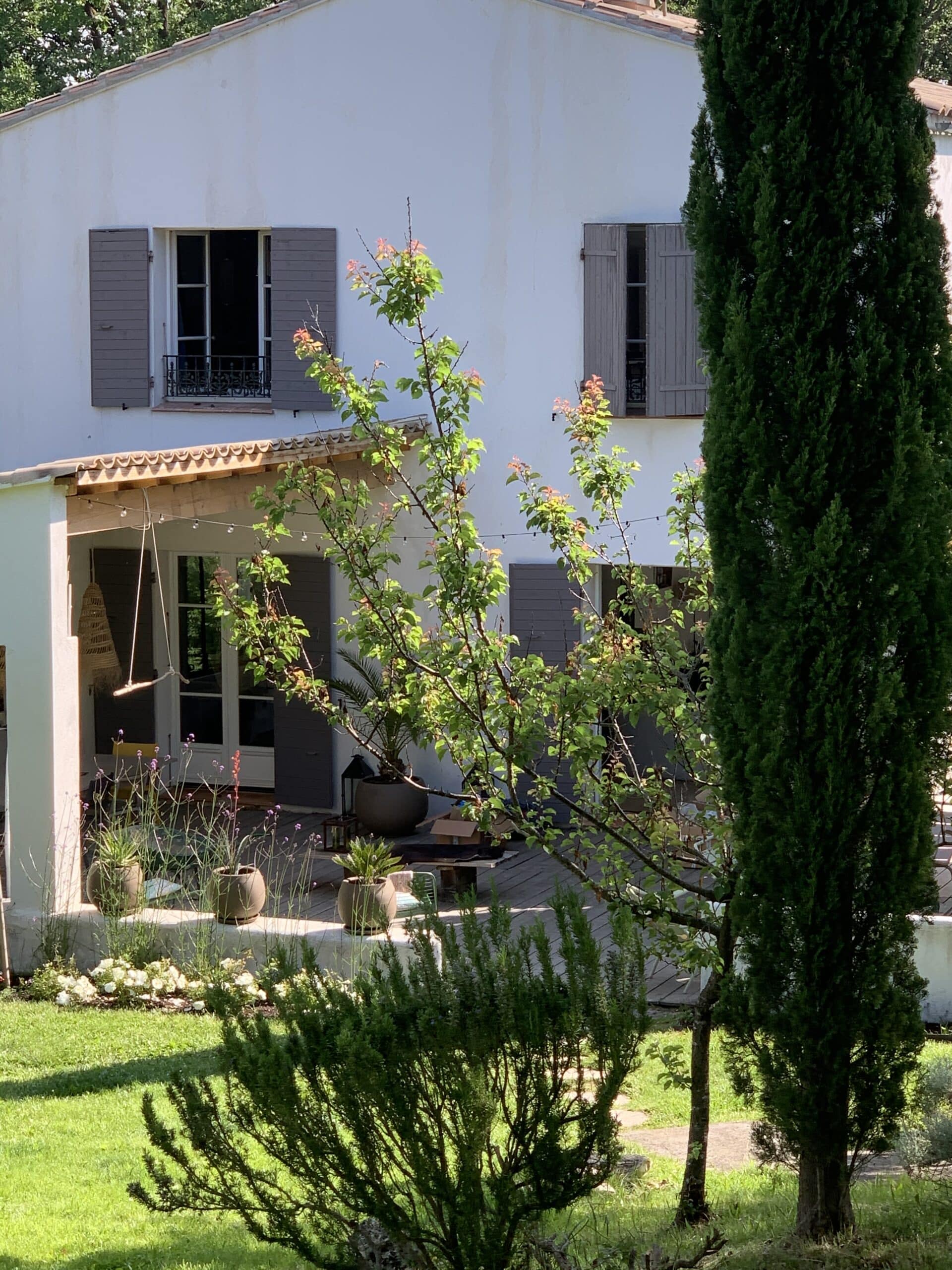 Façade blanche d'une maison ensoleillée avec volets bruns, jardin verdoyant luxuriant et terrasse ombragée, arbres. Panneau: LA BELLE VIE.