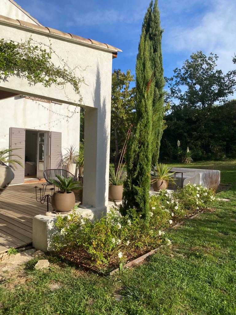 Maison blanche ensoleillée avec terrasse en bois, cyprès élancé et rosiers blancs en fleurs. Des plantes en pot décorent le coin détente extérieur.