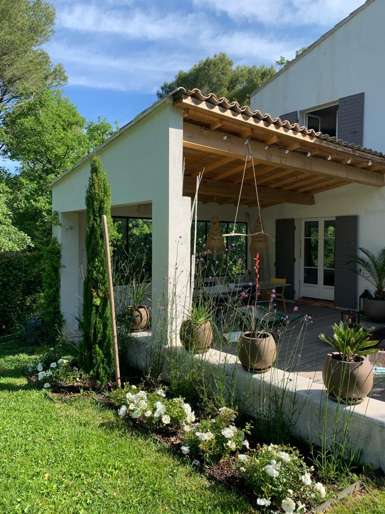 Maison méditerranéenne avec terrasse en bois sous pergola, jardin verdoyant, roses blanches, et ciel bleu ensoleillé.