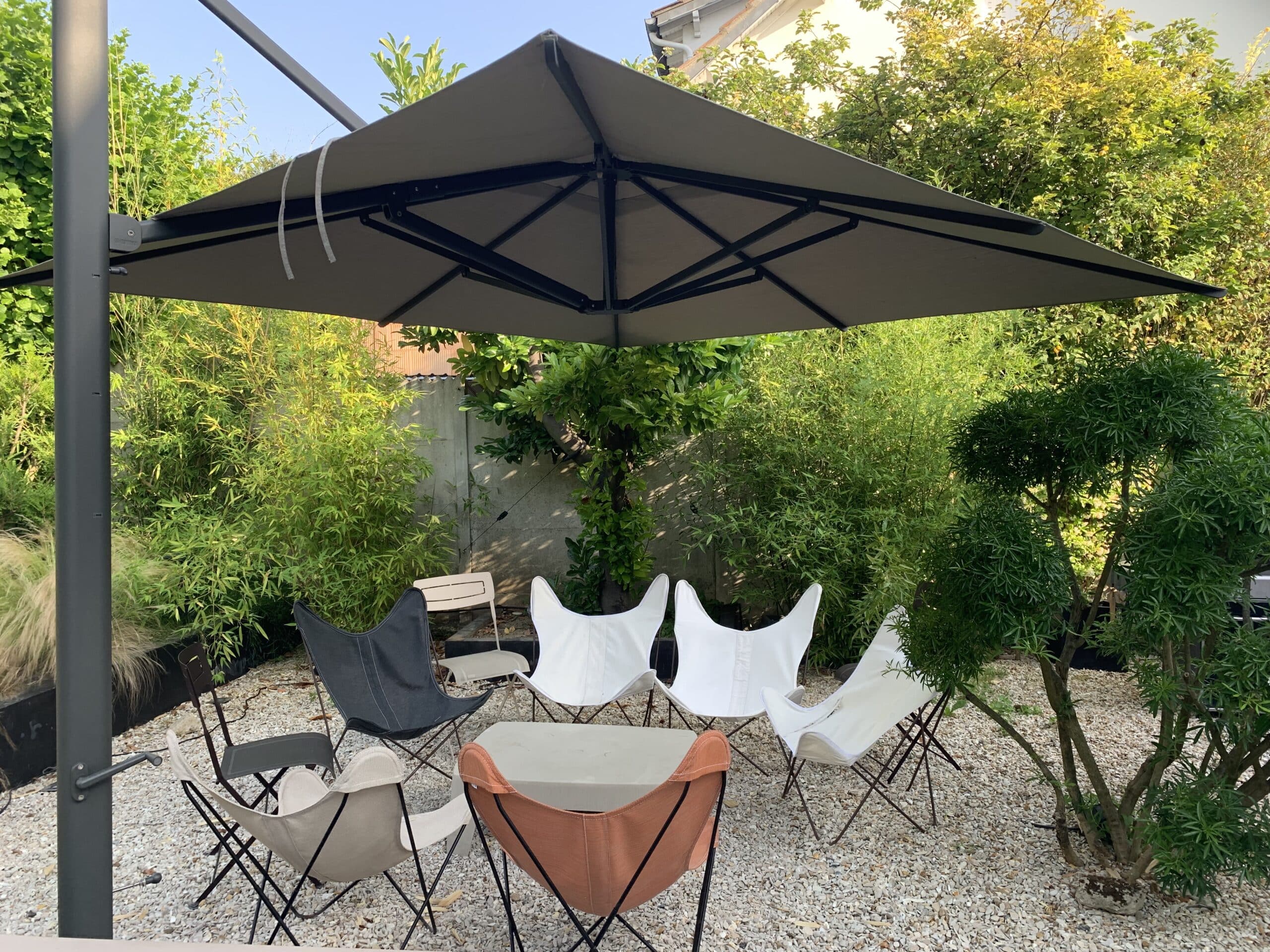 Patio moderne avec grand parasol déporté gris, 6 chaises papillon colorées, table basse sur gravier clair et verdure luxuriante.