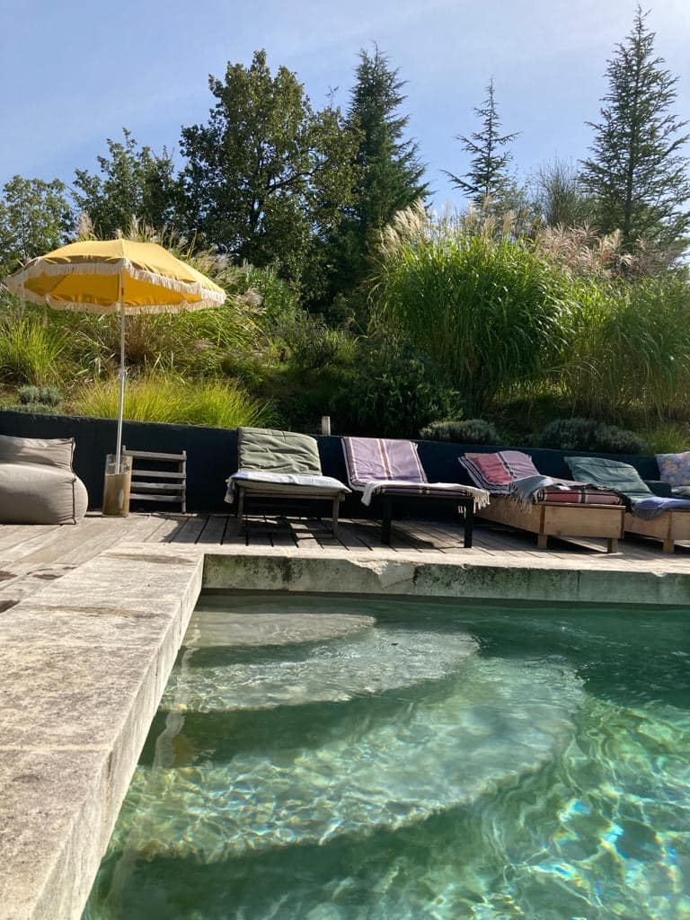 Piscine naturelle en pierre avec marches et terrasse en bois. Chaises longues, parasol jaune. Végétation luxuriante, ciel bleu ensoleillé.