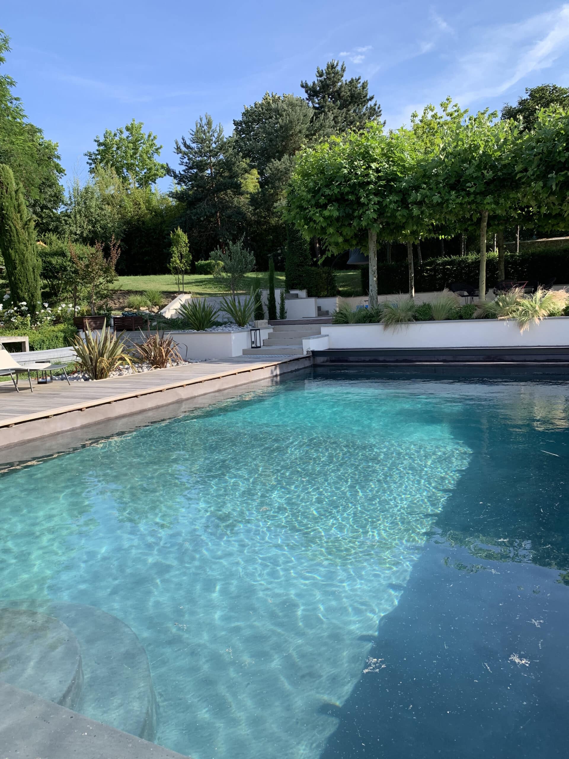 Grande piscine moderne aux eaux turquoise, bordée d'une terrasse en bois. Jardin luxuriant, arbres verts, escaliers et mobilier extérieur.