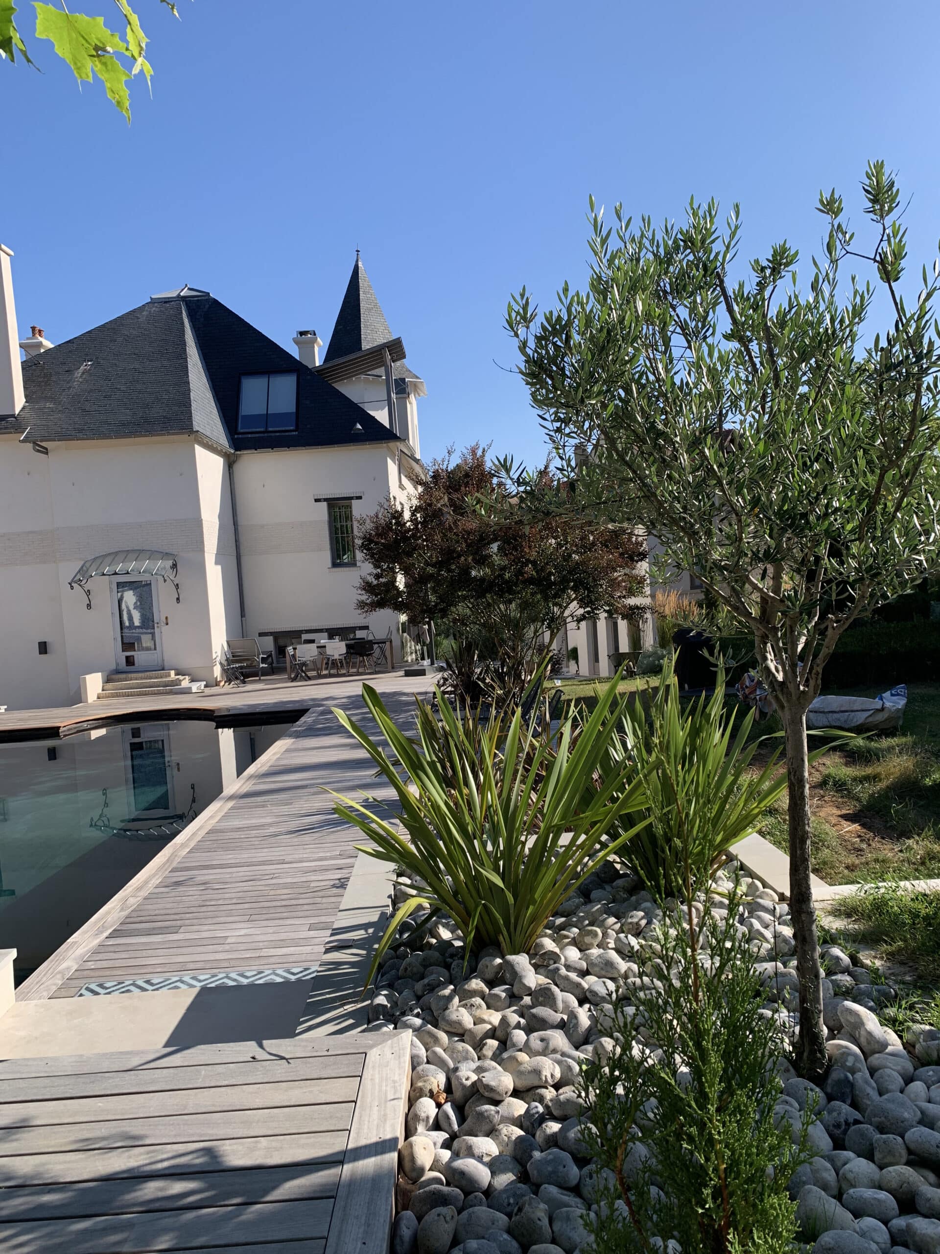 Grande maison blanche avec toit noir, piscine miroir et terrasse bois. Jardin méditerranéen avec galets et olivier ensoleillé.