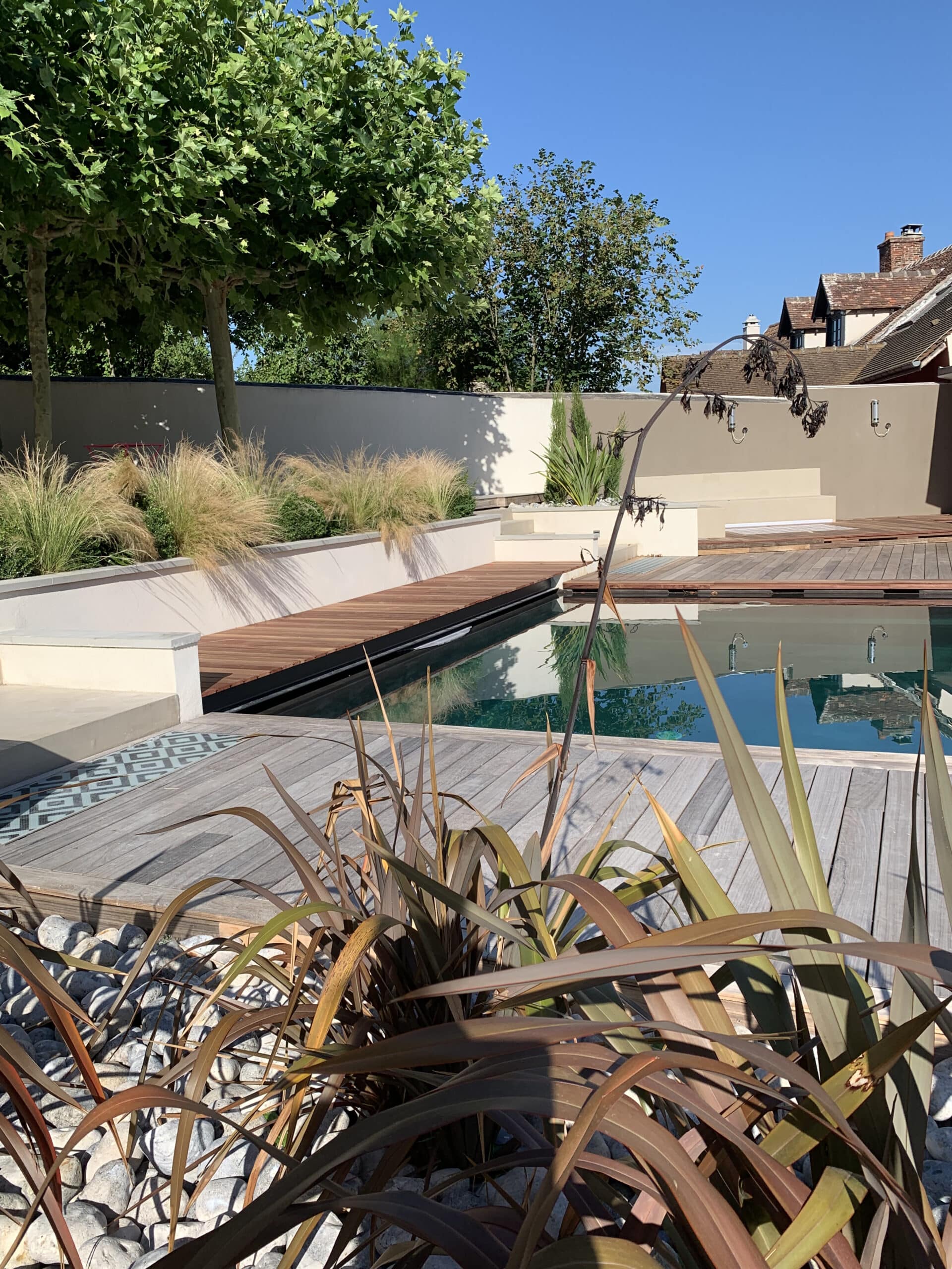 Jardin moderne: piscine, terrasse bois, plantes en bacs, graminées, galets. Murs clairs, arbres et maison ancienne sous ciel bleu.