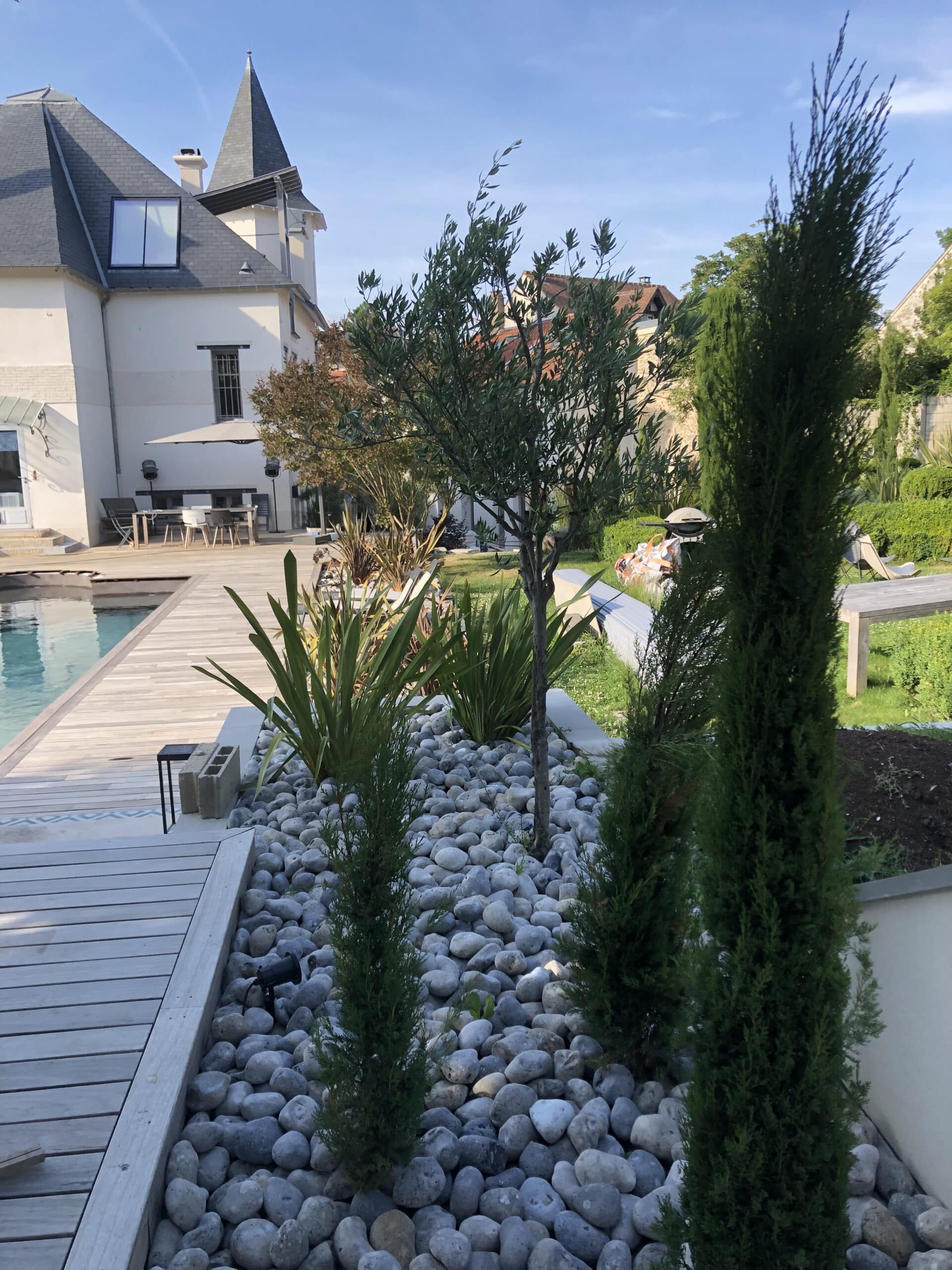 Piscine azur et terrasse en bois clair. Jardin paysagé zen avec galets blancs, olivier et plantes. Grande maison élégante sous ciel bleu.