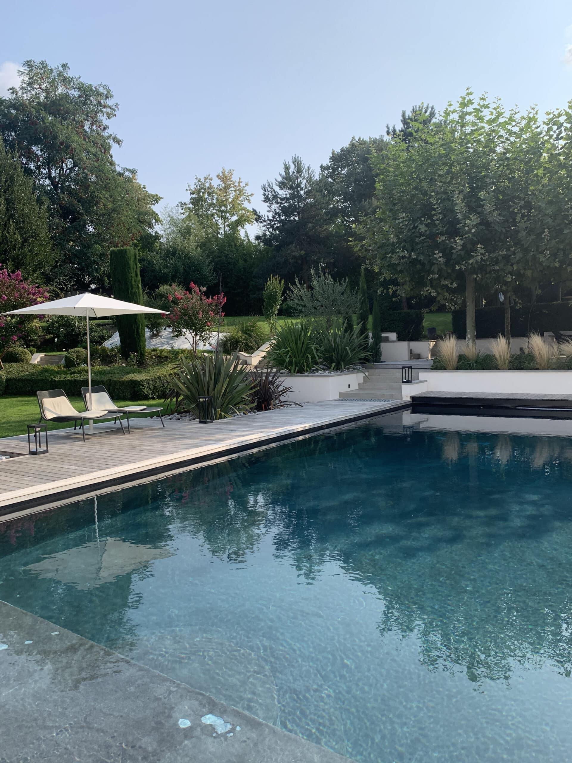 Piscine moderne bleu-vert avec terrasse en bois, deux chaises longues et un parasol blanc. Grand jardin luxuriant et ciel clair.