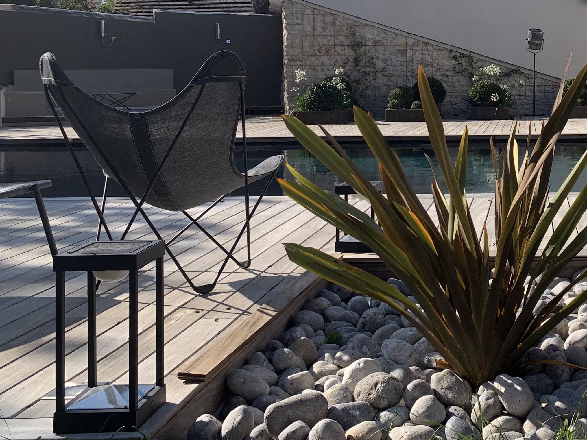 Fauteuil papillon gris, lanterne solaire sur terrasse en bois bordant une piscine. Rochers, plante exotique et maison moderne en arrière-plan.