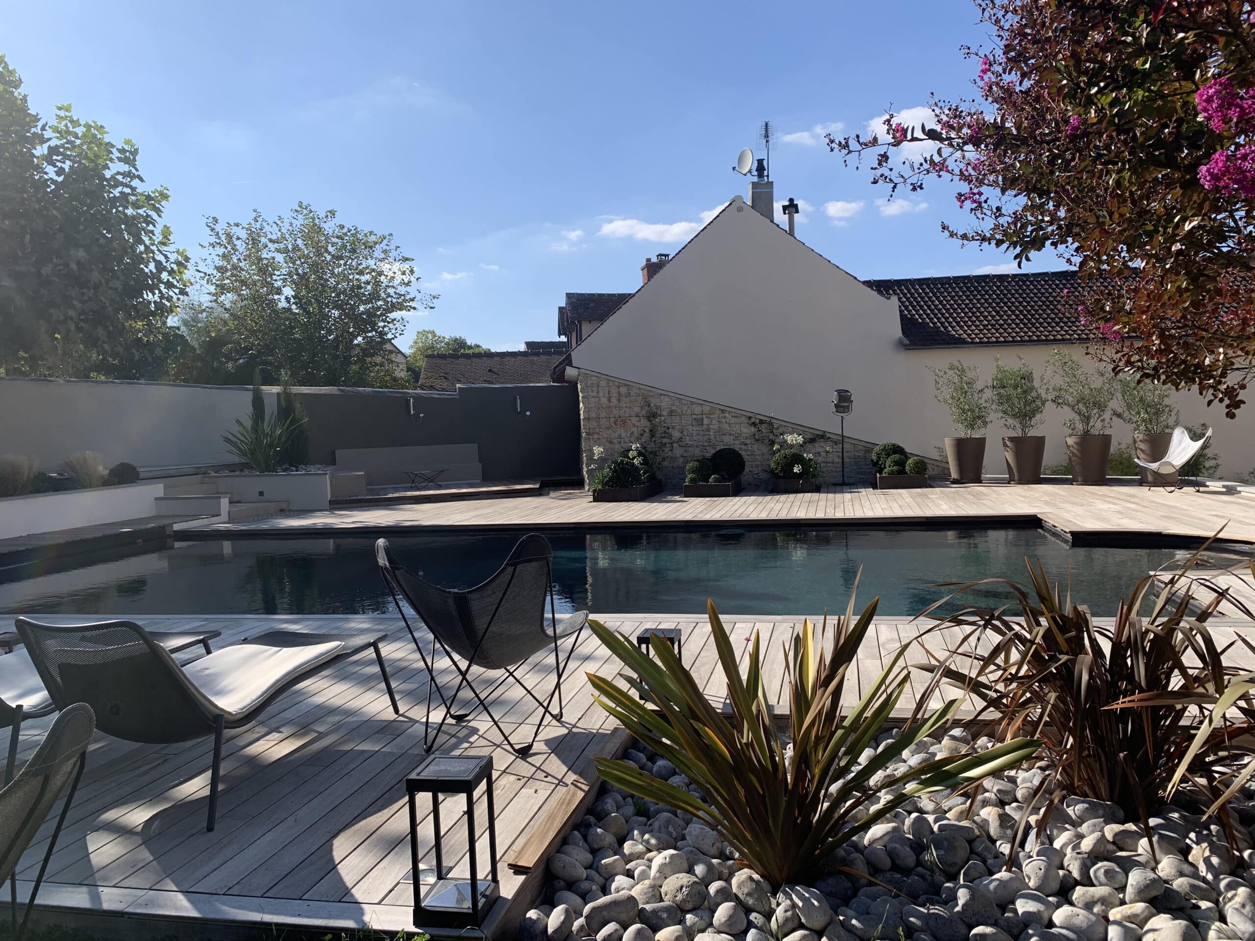 Grande piscine sombre avec terrasse bois, chaises modernes, plantes et murs d'une maison contemporaine sous ciel bleu ensoleillé.