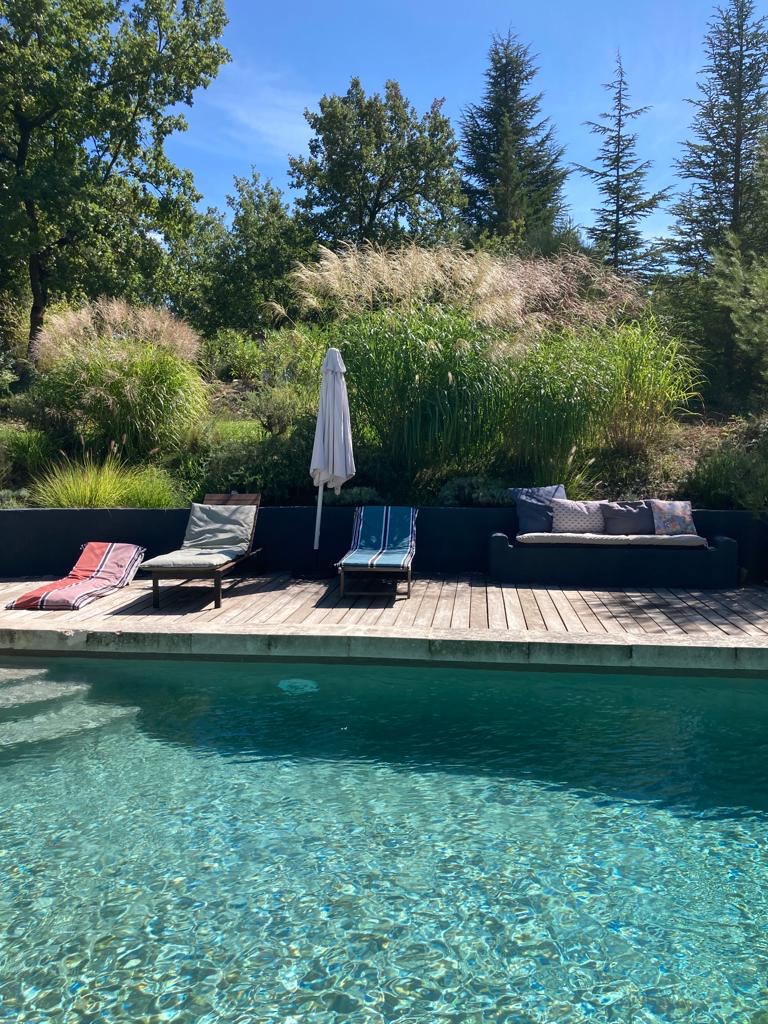 Piscine moderne avec terrasse en bois, transats, canapé extérieur et jardin luxuriant sous un ciel bleu ensoleillé.