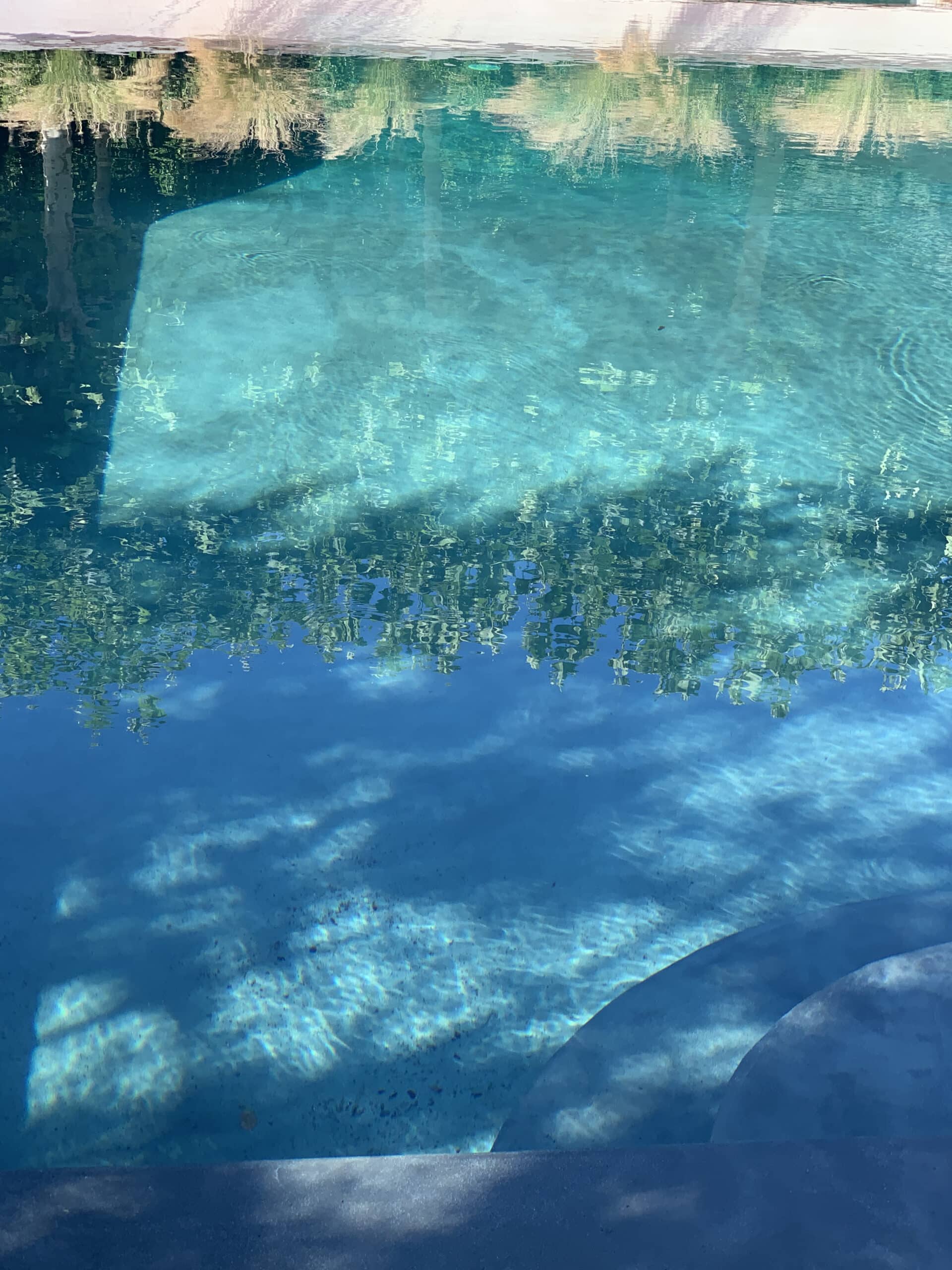 Eau turquoise d'une piscine ensoleillée, avec des reflets ondulants de ciel et de verdure. Le fond clair et des marches incurvées sont visibles.