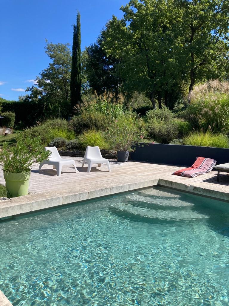 Jardin luxuriant avec piscine turquoise, terrasse en bois, deux transats blancs et un drap rayé. Journée ensoleillée parfaite pour la détente.