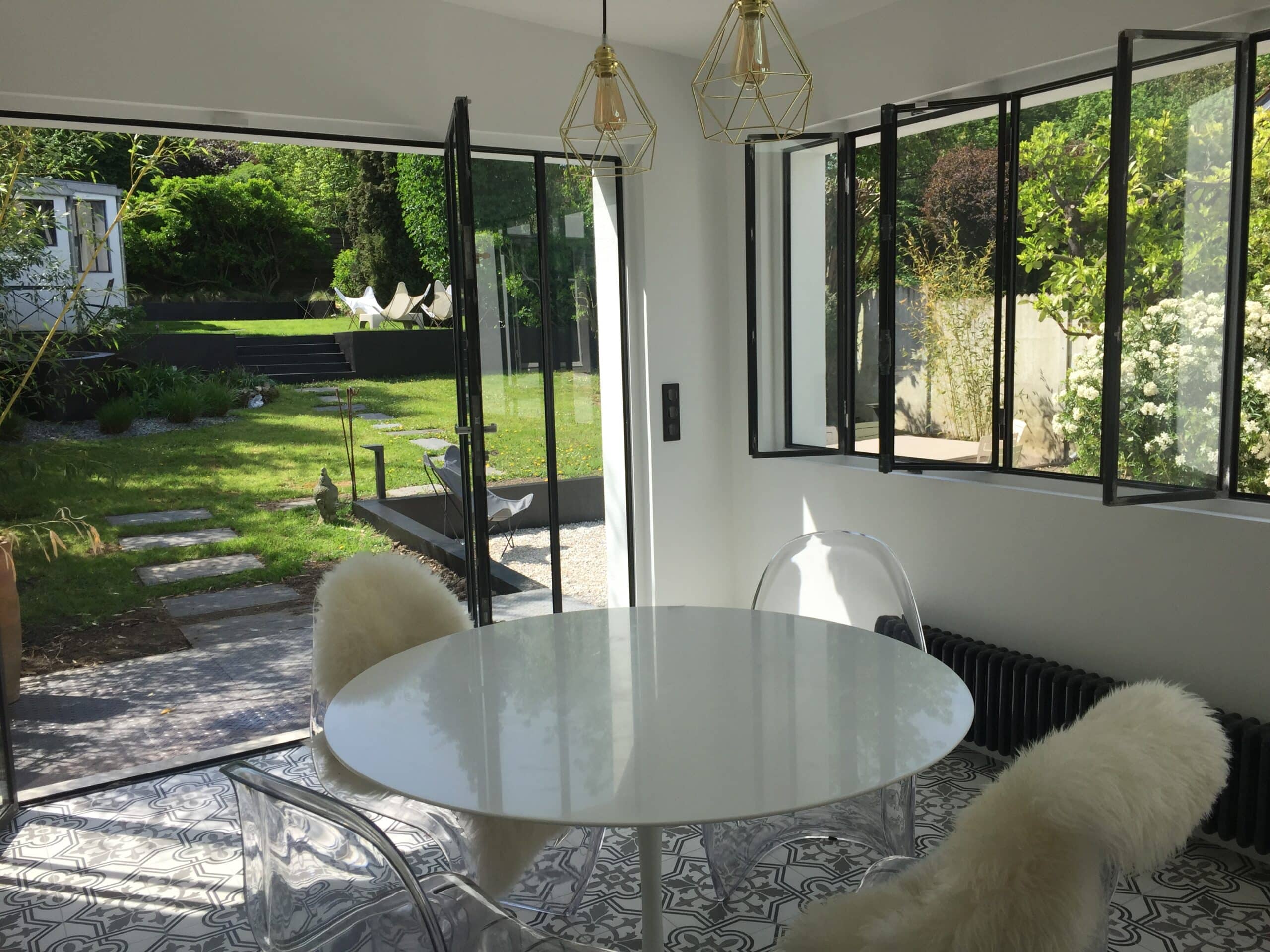 Salle à manger moderne lumineuse avec table ronde blanche, chaises transparentes et vue sur un jardin verdoyant. Baies vitrées noires et sol à motifs.