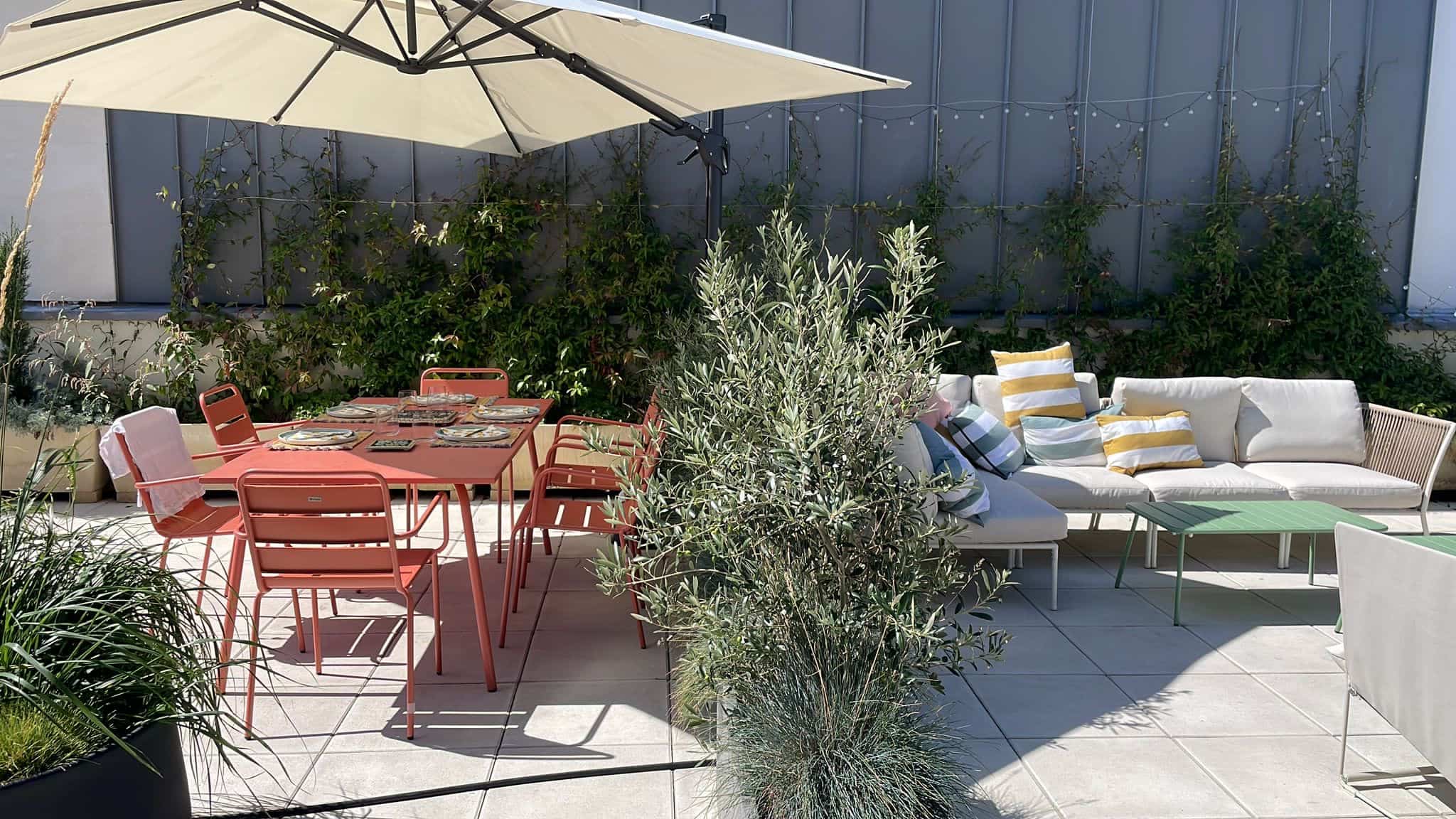 Grande terrasse ensoleillée avec table à manger corail sous parasol, salon de jardin confortable et plantes méditerranéennes.