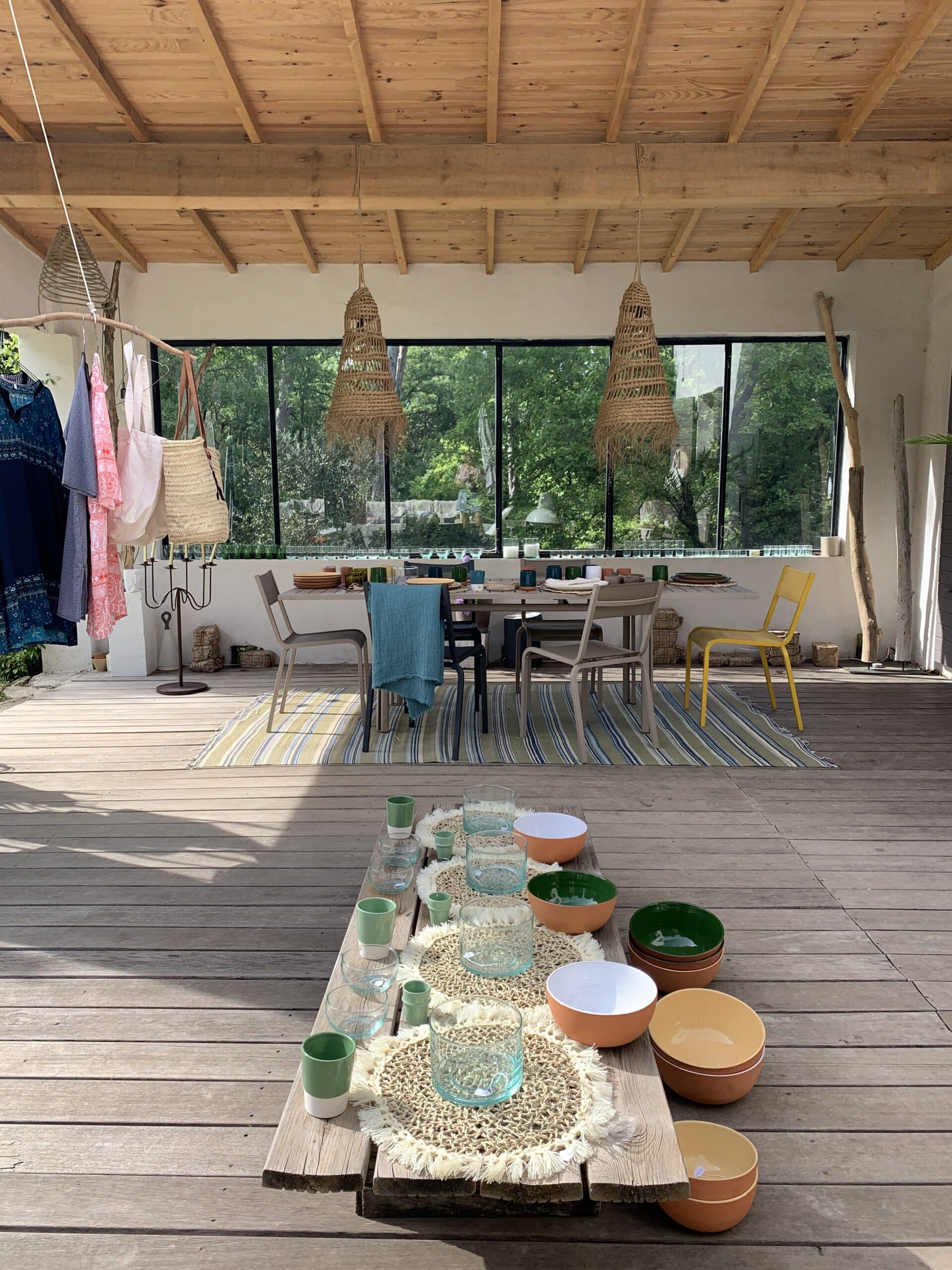 Terrasse bois avec table dressée, vaisselle artisanale colorée, gobelets en verre. Vêtements, sacs, lampes tressées. Vue nature luxuriante.