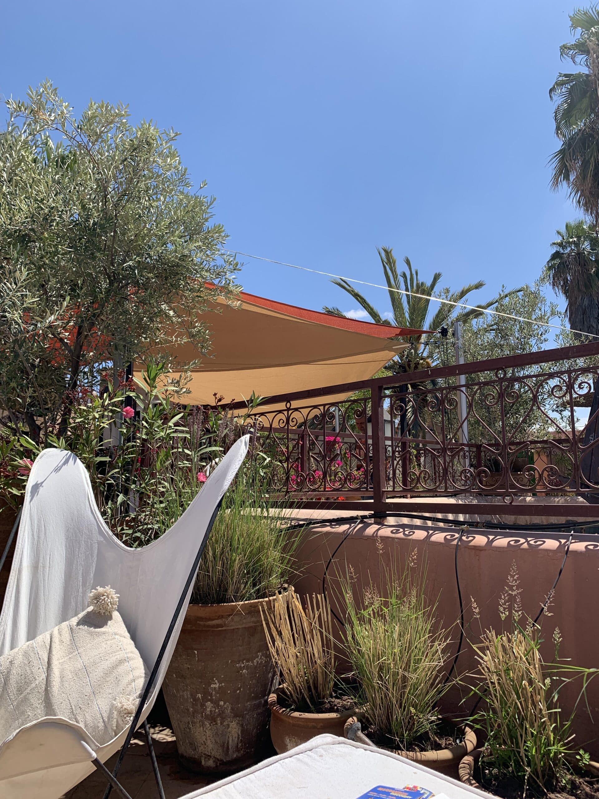 Terrasse ensoleillée avec fauteuil papillon blanc, coussin à pompons, plantes en pot, balustrade ornée et voiles d'ombrage. Ciel bleu, oliviers et palmiers.