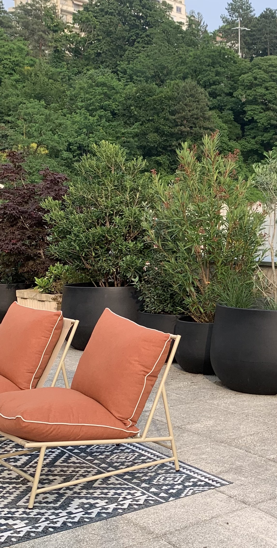 Deux chaises d'extérieur orange sur un tapis géométrique, entourées de grands pots noirs avec plantes vertes, sur une terrasse urbaine.