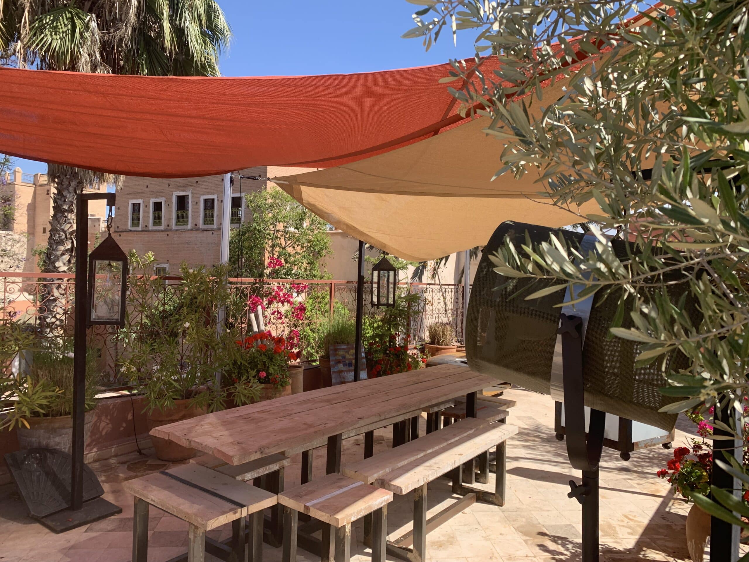 Terrasse ensoleillée style méditerranéen avec table et bancs en bois, voiles d'ombrage orange et beige, plantes fleuries, palmier et lanterne. Vue paisible.