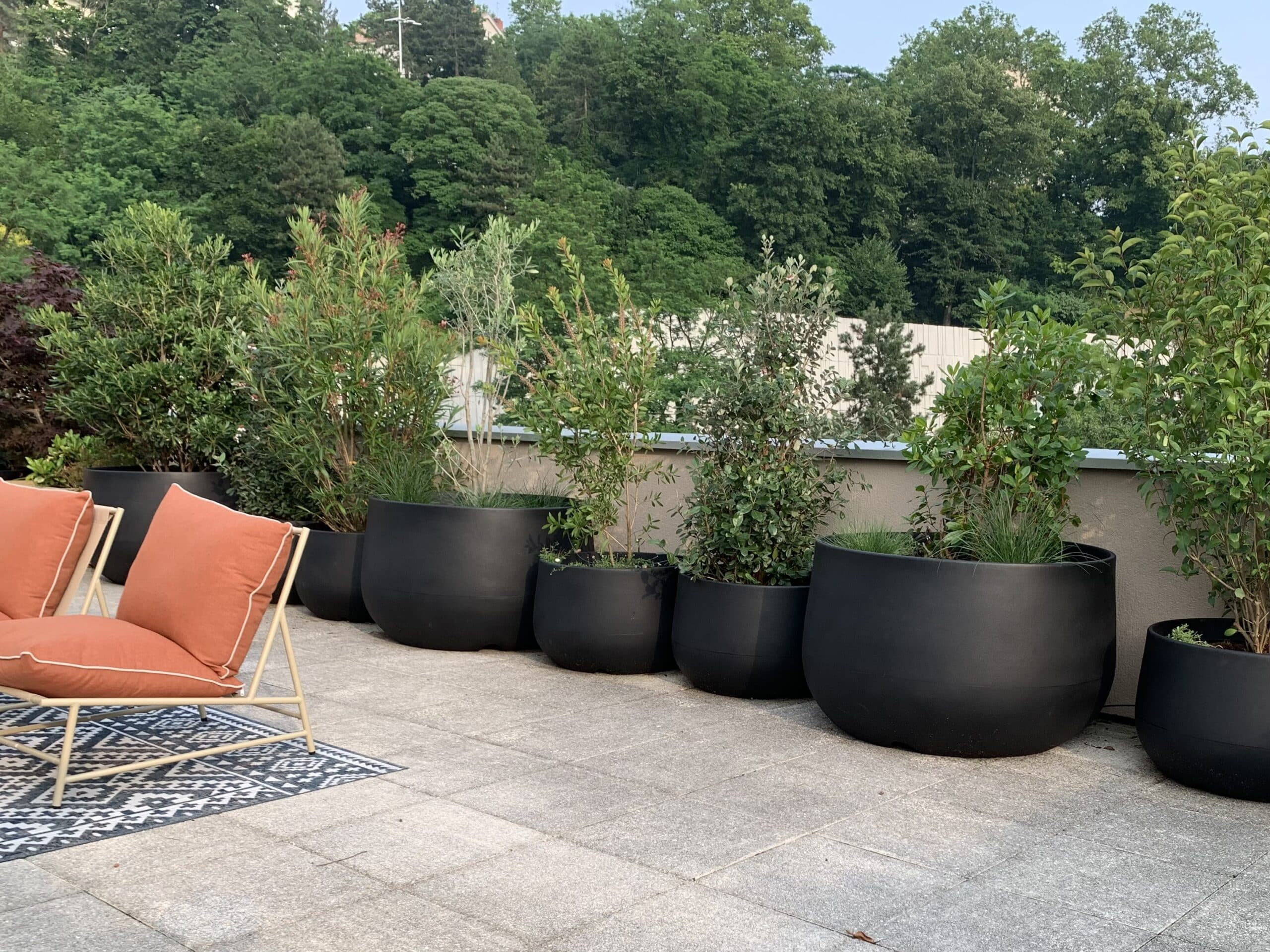 Terrasse moderne: jardinières noires avec plantes vertes, fauteuils orange, tapis géométrique sur patio gris, fond de verdure luxuriante.