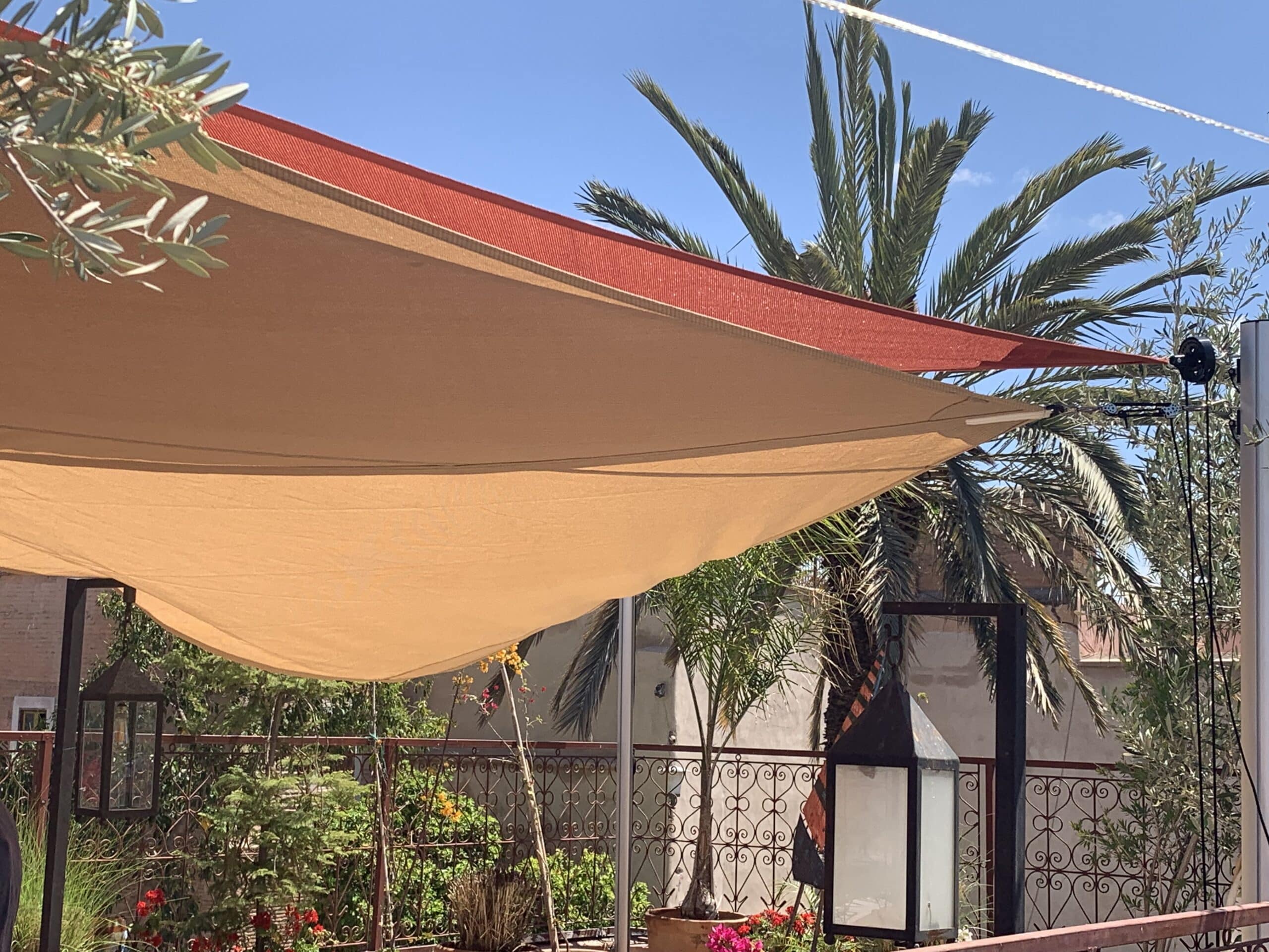 Terrasse ensoleillée avec voile d'ombrage orange et rouge, palmiers, verdure luxuriante et clôture en fer forgé sous un ciel bleu clair.