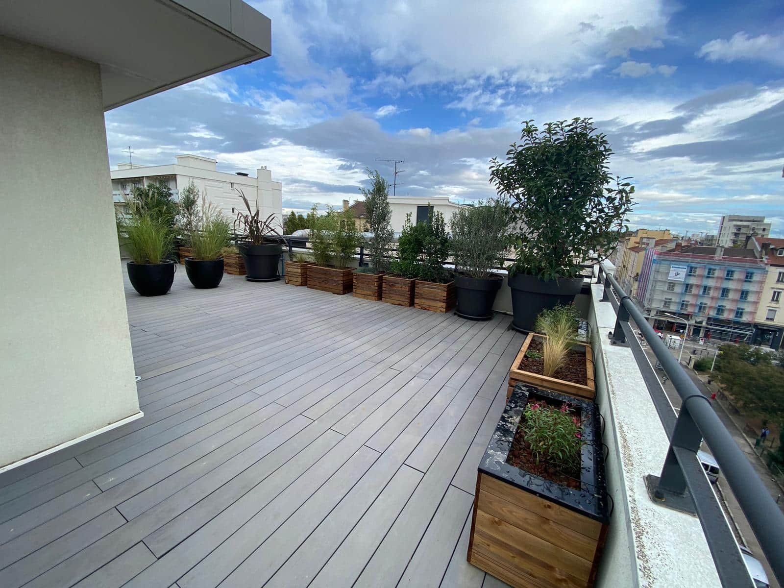 Terrasse de toit moderne avec sol en decking gris, bordée de grandes jardinières de plantes et vue sur la ville urbaine ensoleillée.