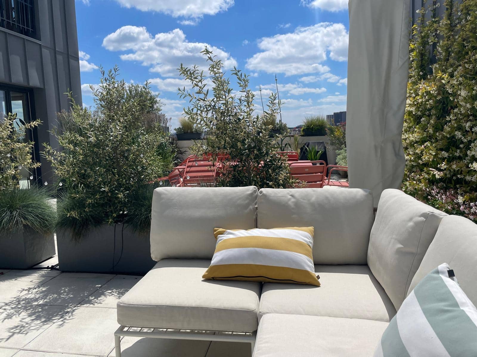 Terrasse sur le toit ensoleillée: canapé d'extérieur beige avec coussin rayé, plantes luxuriantes et chaises rouges sous ciel bleu. Vue relaxante.