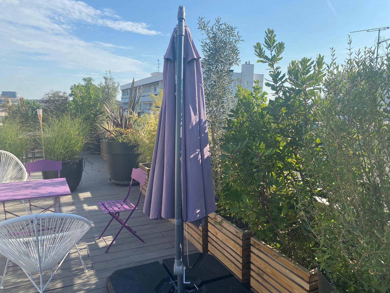 Terrasse sur le toit avec mobilier violet et blanc, parasol fermé, grands pots de plantes vertes et arbustes sous un ciel bleu dégagé.