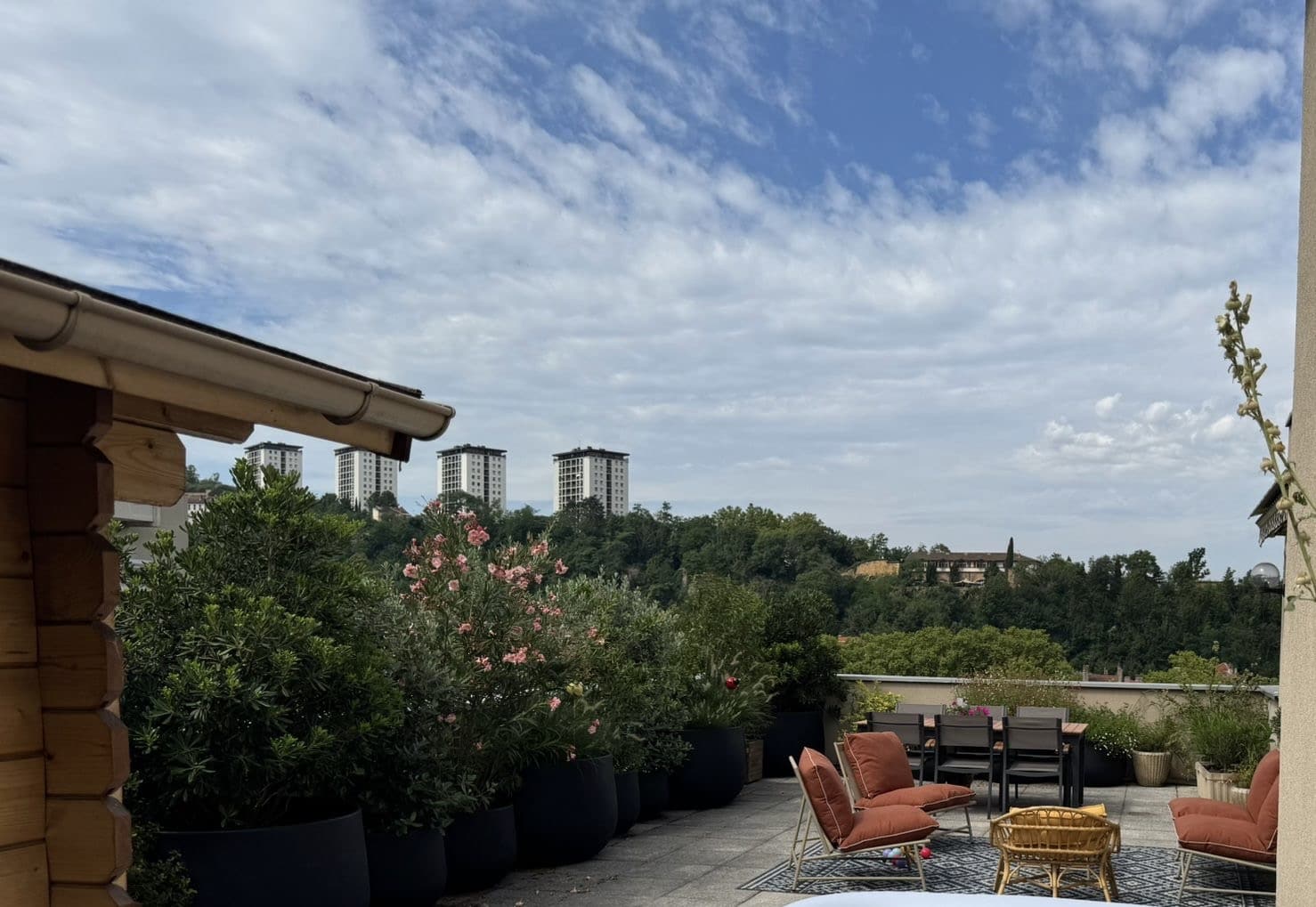 Terrasse de toit aménagée avec mobilier extérieur et plantes en pots, vue sur des immeubles blancs au loin dominant une colline boisée, ciel bleu et nuageux.