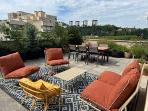 Terrasse de toit aménagée avec salon extérieur couleur terracotta, tapis à motifs, table basse, et vue sur des immeubles et une colline boisée.