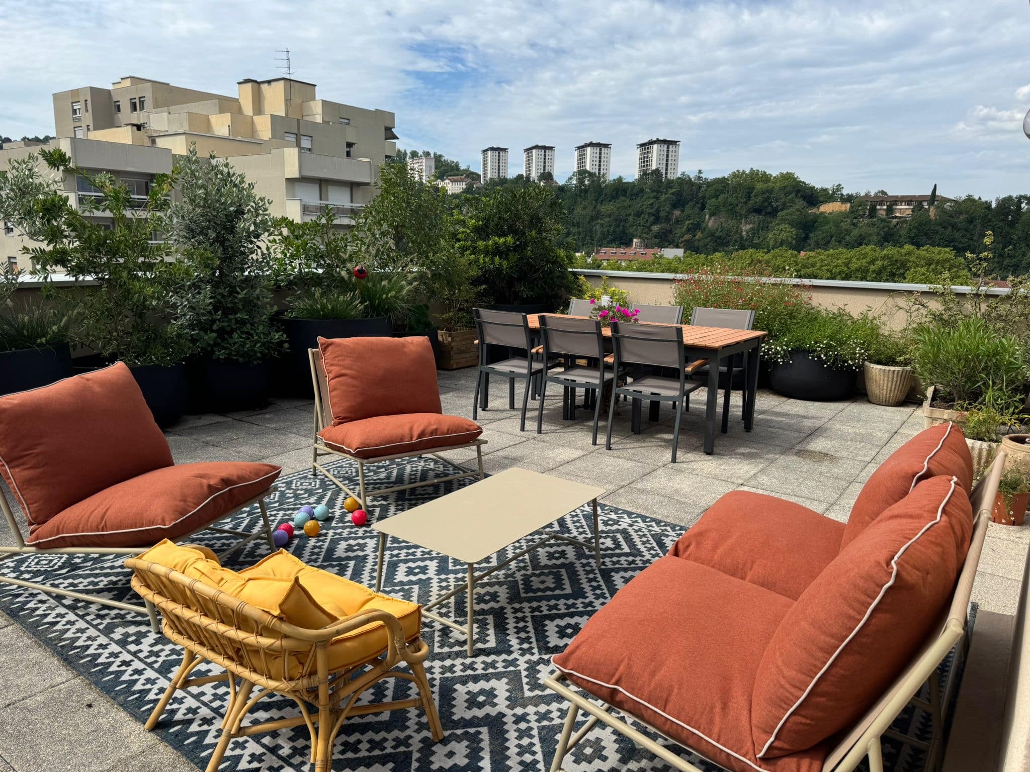 Terrasse de toit aménagée avec salon extérieur couleur terracotta, tapis à motifs, table basse, et vue sur des immeubles et une colline boisée.