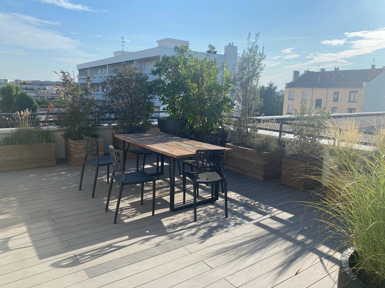 Terrasse de toit ensoleillée avec table en bois, chaises sombres et plantes vertes en jardinières sur un decking clair, avec des bâtiments en fond.