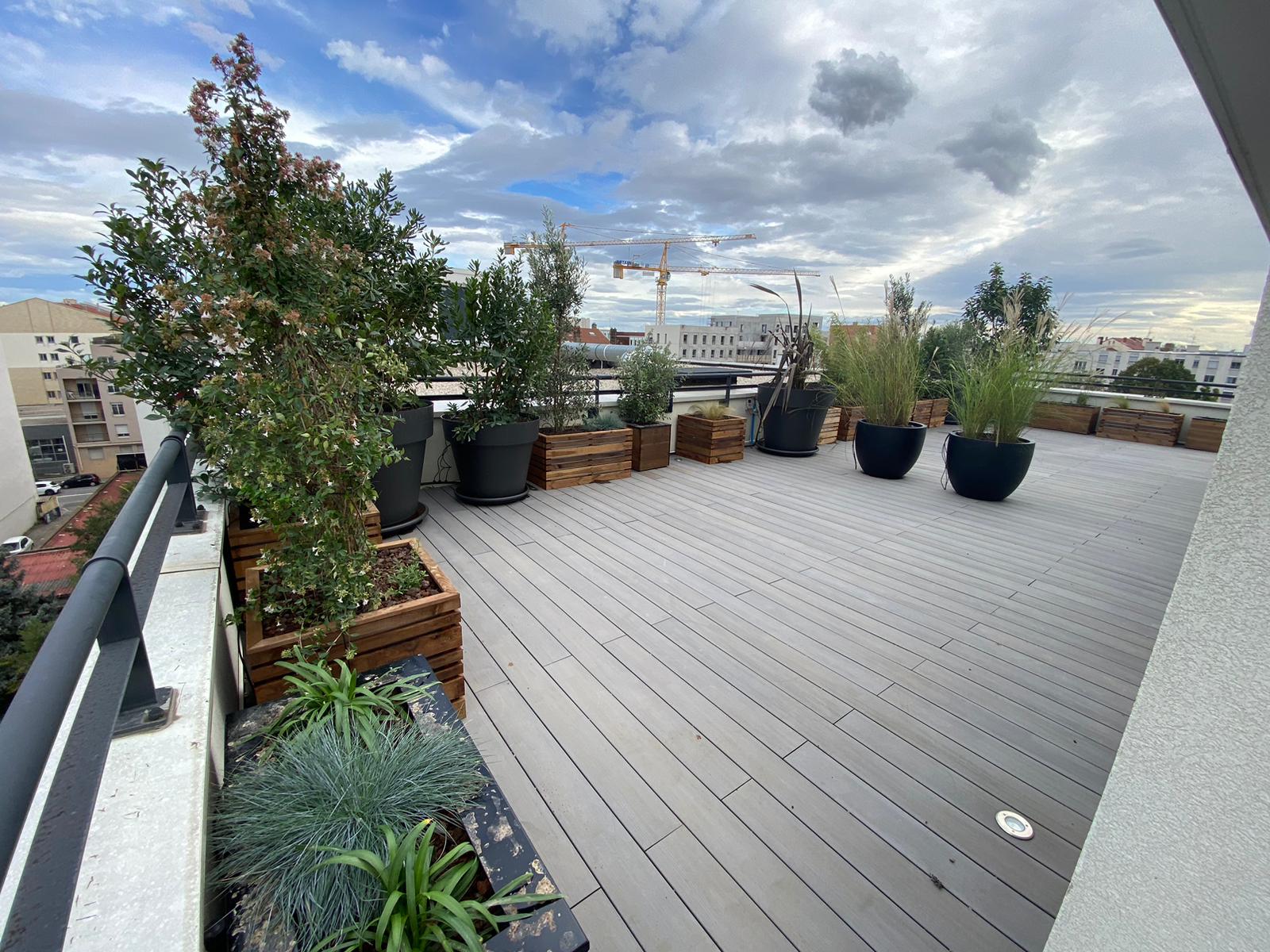 Terrasse sur le toit avec decking clair, plantes variées en pots et jardinières bois. Vue urbaine, grues et ciel nuageux.