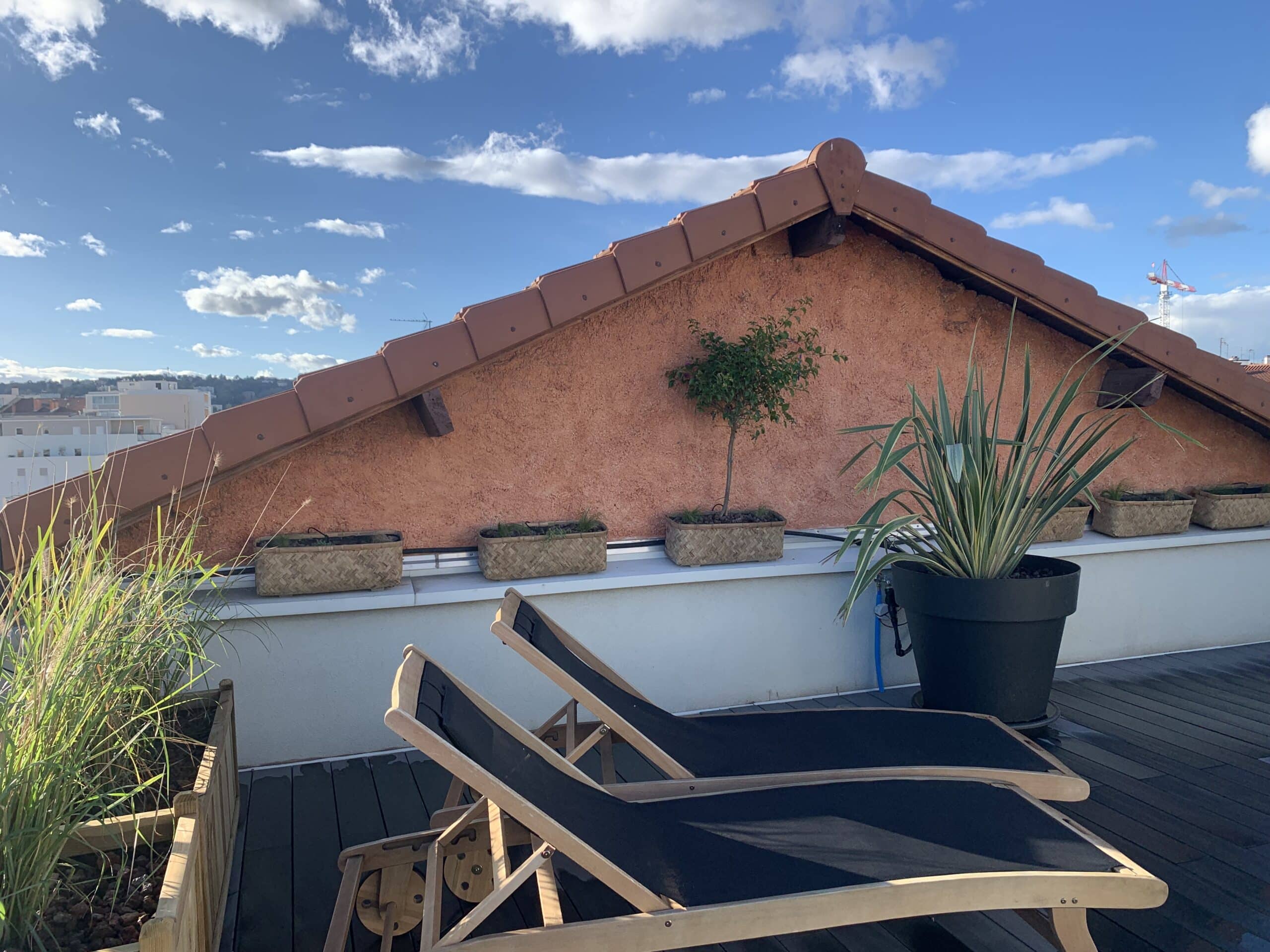 Terrasse ensoleillée avec deux chaises longues noires sur un platelage bois. Verdure luxuriante, mur ocre, toit tuilé et ciel bleu nuageux.